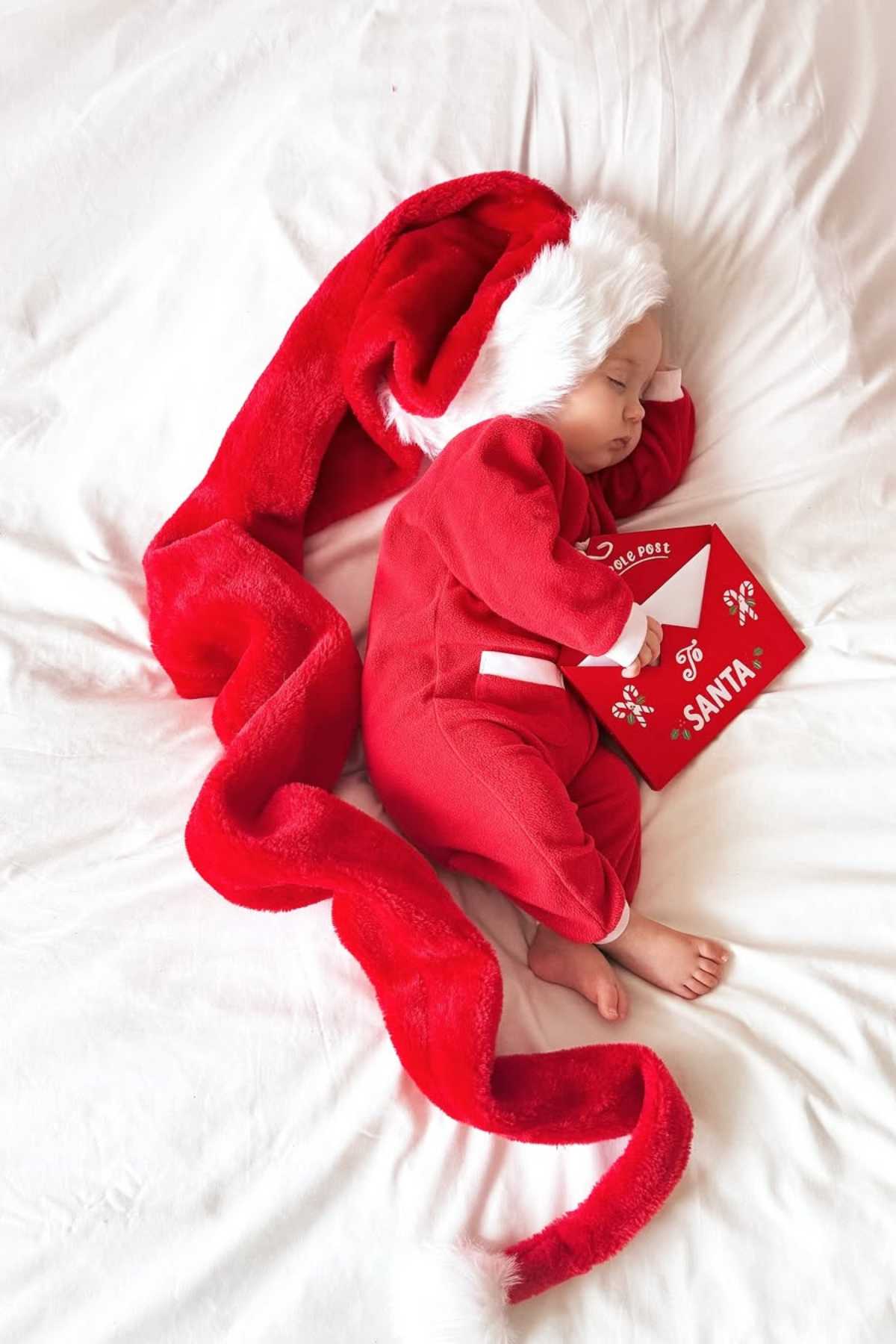 Baby wearing a long red Santa hat and red pajamas sleeps on a white bed while holding a red envelope addressed to Santa.
