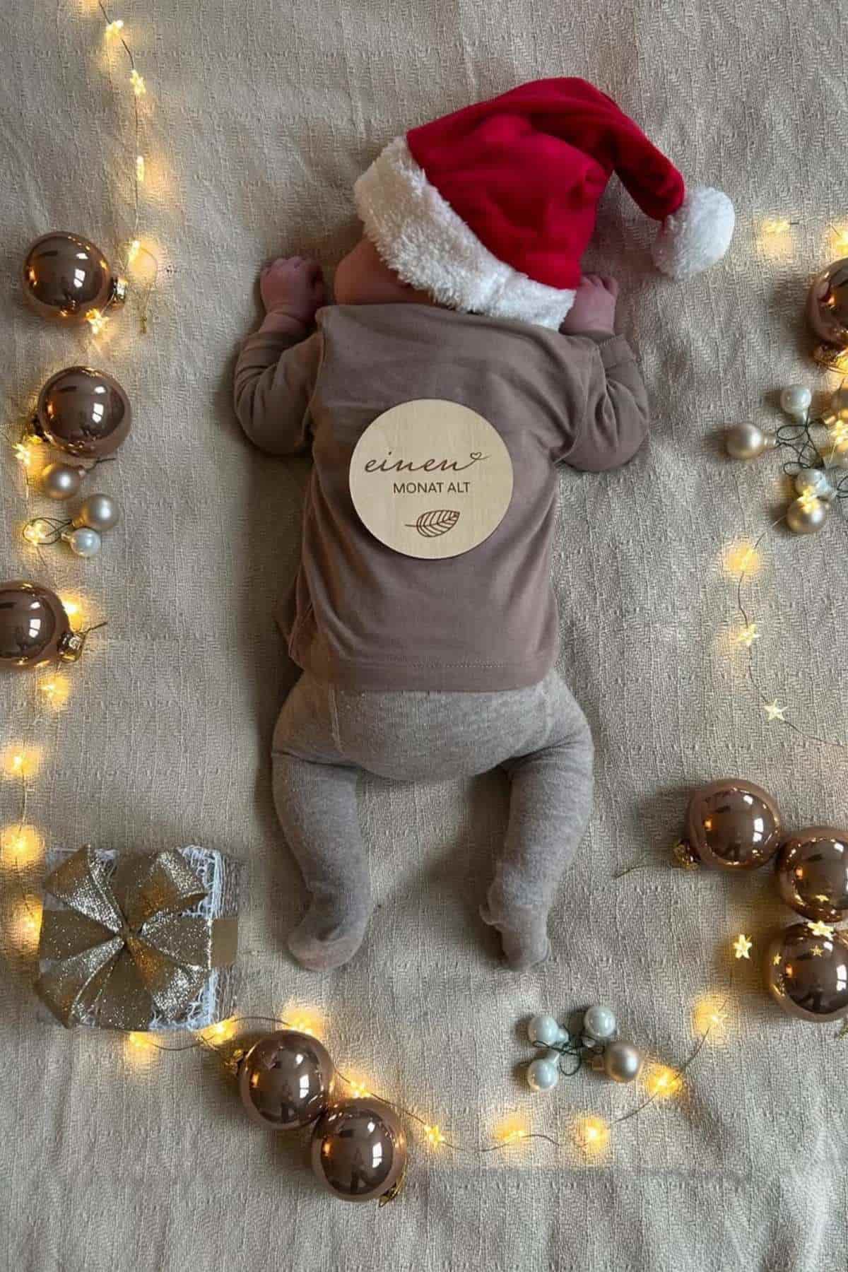 A baby in a Santa hat lies on a blanket surrounded by festive ornaments and string lights, with a sign on their back reading "einen MONAT ALT.