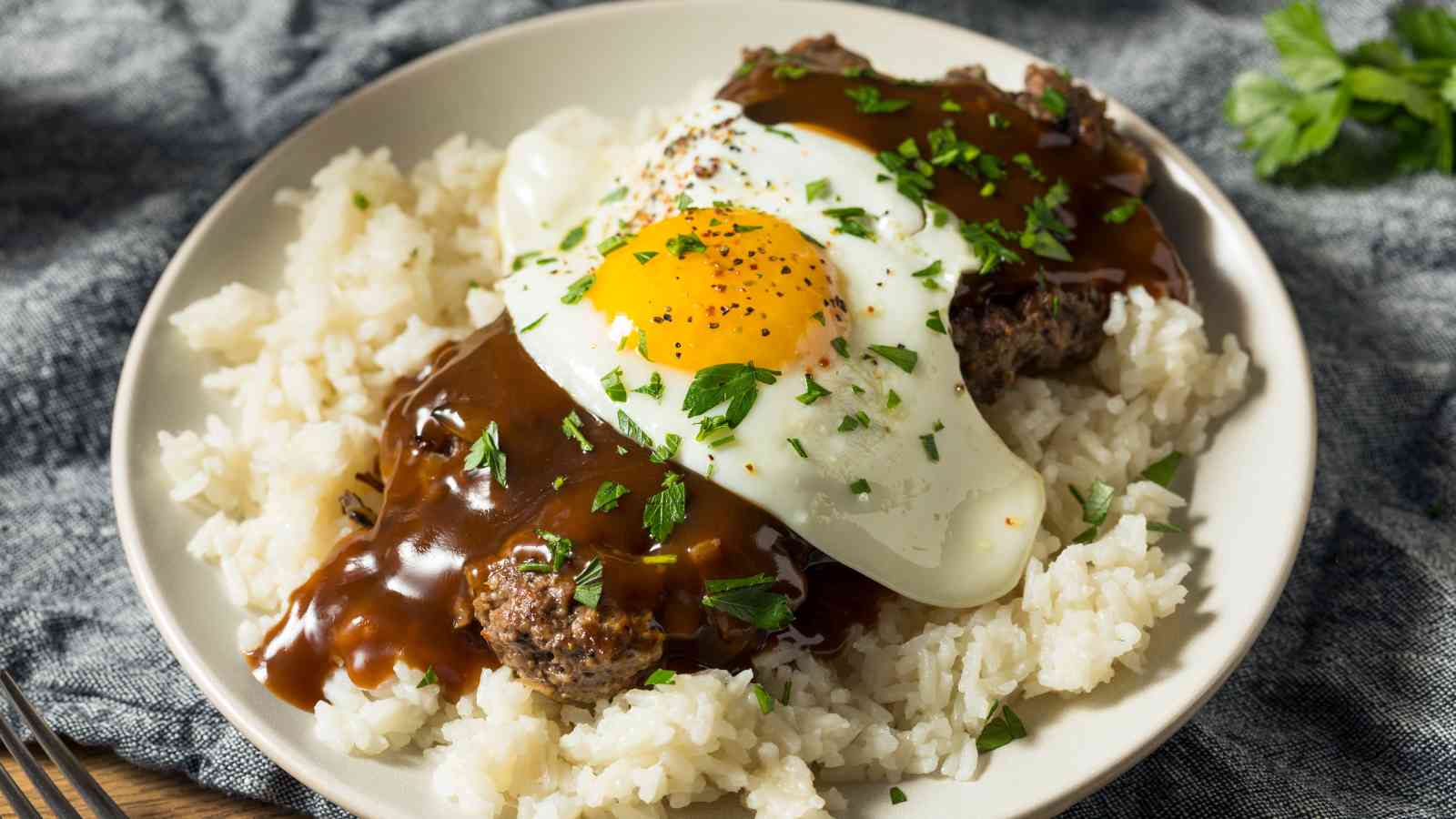 A plate of white rice topped with a hamburger patty, brown gravy, a sunny-side-up egg, and chopped herbs.
