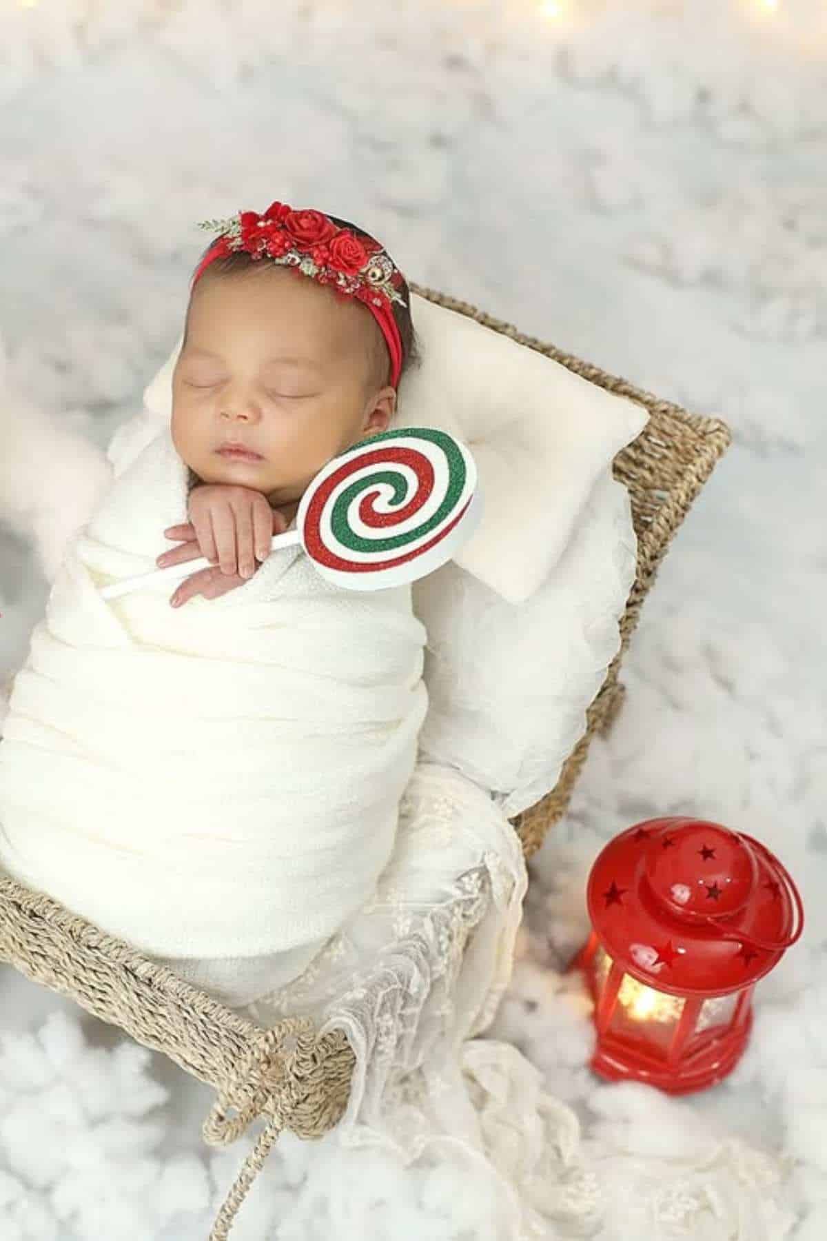 A newborn baby wrapped in a white blanket lies in a basket, holding a red, green, and white lollipop, with a red lantern nearby on a snowy white background.