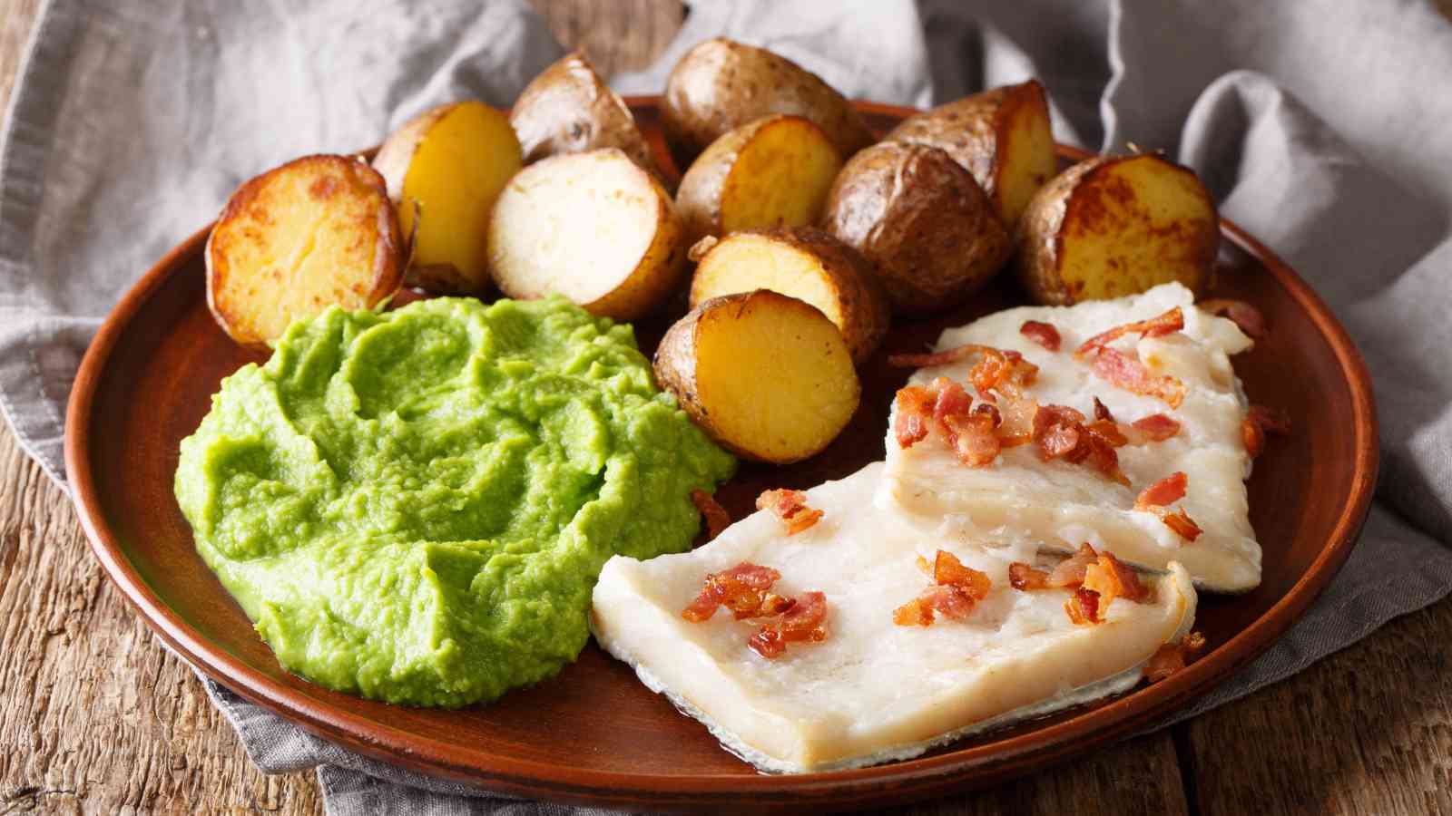 A plate with two pieces of white fish topped with bacon, mashed peas, and roasted potato halves, on a wooden table with a gray napkin.