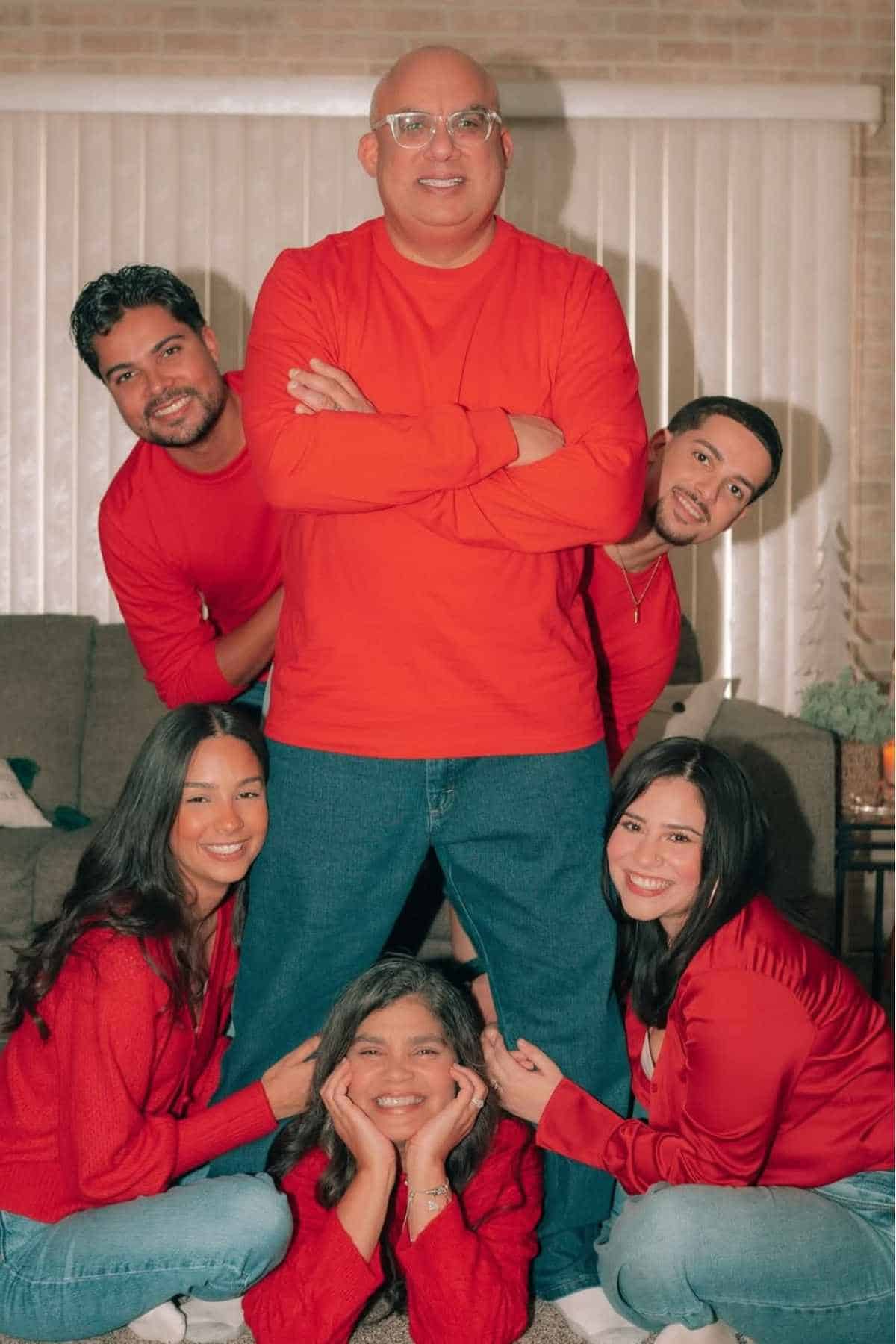 Six people pose indoors, all wearing red tops and blue jeans. Four are sitting or kneeling on the floor, while two stand behind a man with his arms crossed. Everyone is smiling.