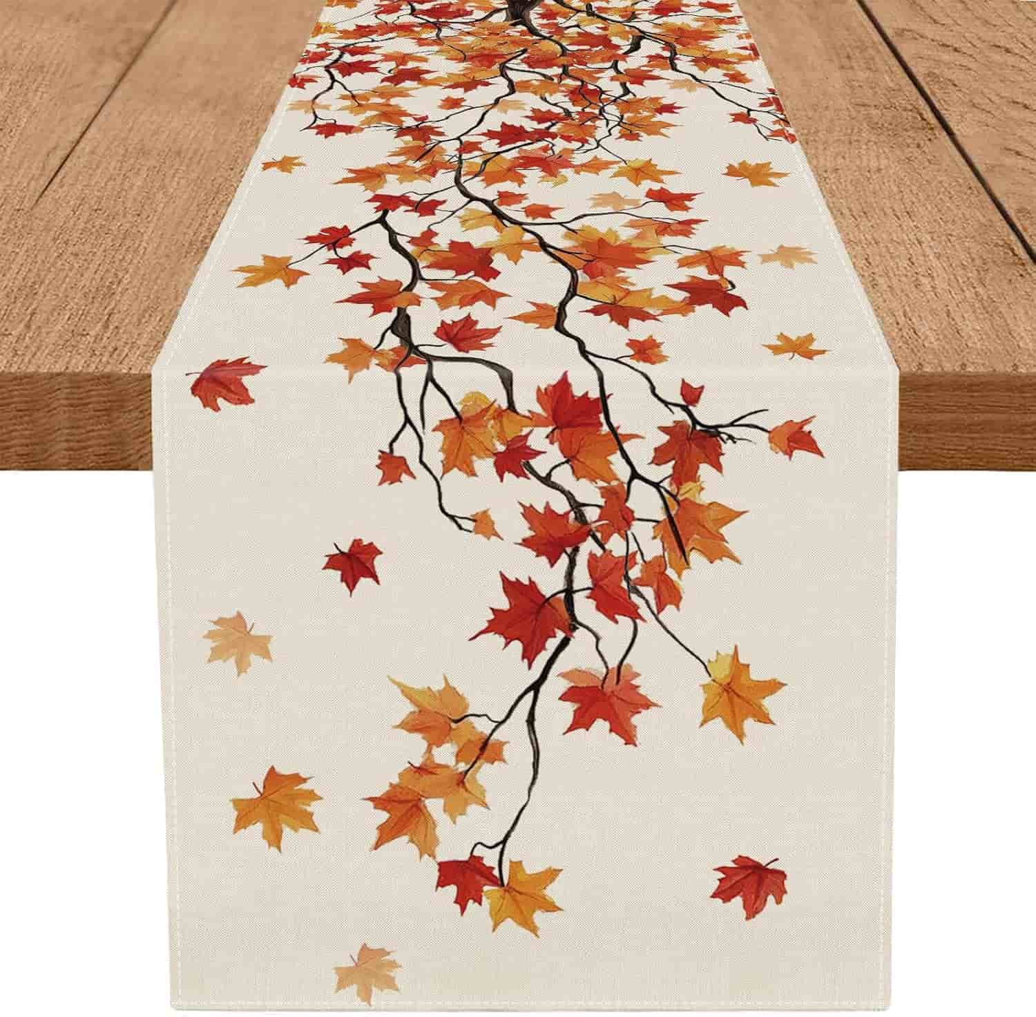 A wooden table with a cream-colored runner featuring a pattern of red, orange, and yellow autumn leaves on black branches.