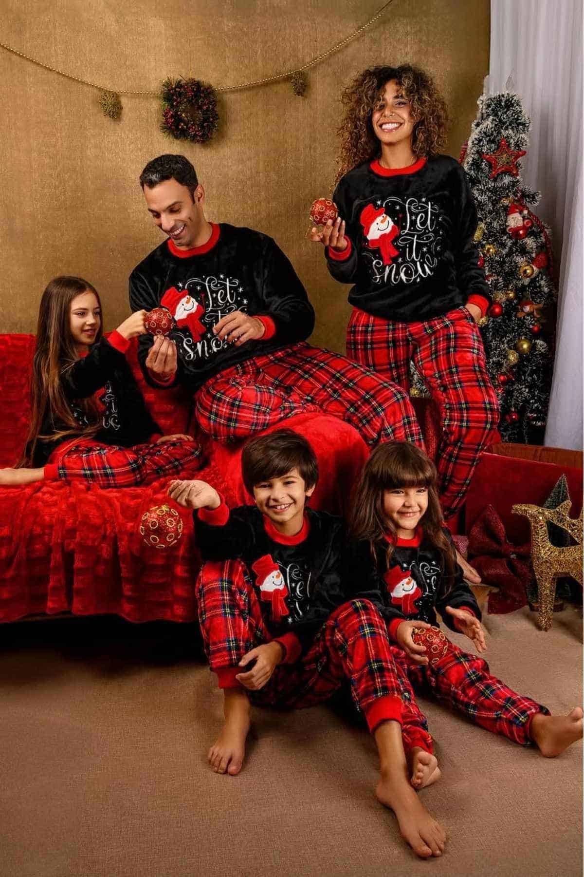 A family of five in matching festive pajamas poses indoors with Christmas decorations and a Christmas tree.
