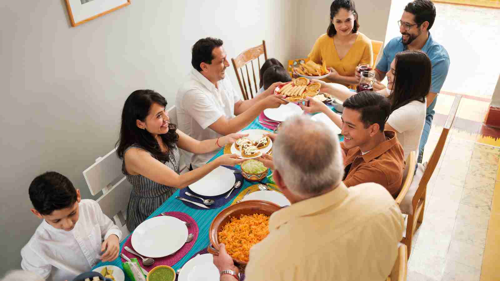 A group of people sit around a dining table, passing and sharing dishes of food, preparing to eat together in a brightly lit room.