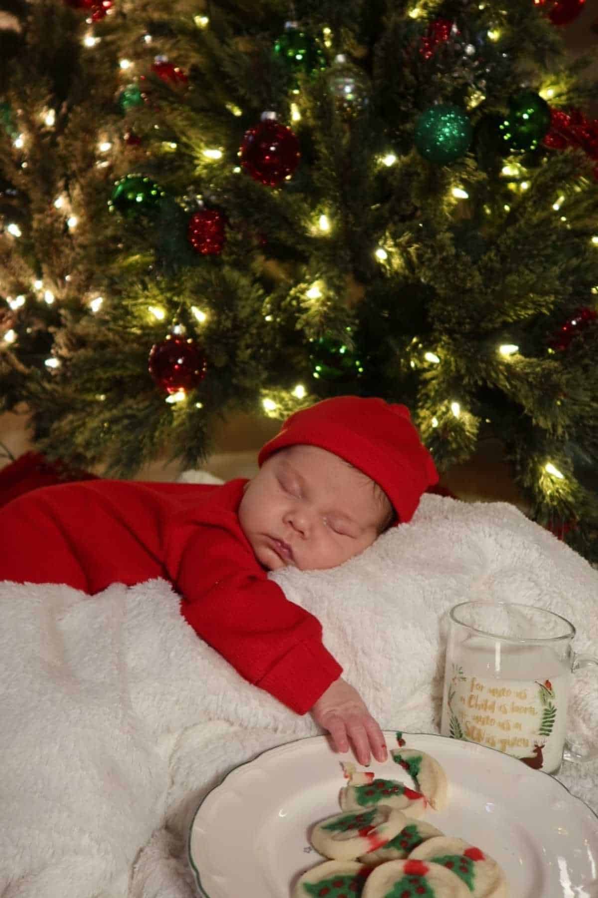 A baby in a red outfit sleeps on a white blanket in front of a decorated Christmas tree, with a plate of cookies and a glass of milk nearby.