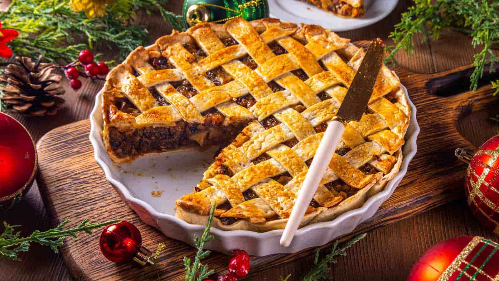 A lattice-topped pie with a slice cut out sits in a white dish on a wooden board, surrounded by festive holiday decorations and greenery.