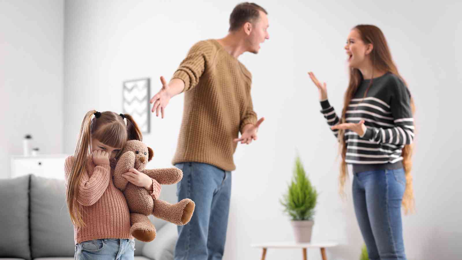 A young girl holding a teddy bear covers her face while two adults argue in the background inside a living room.