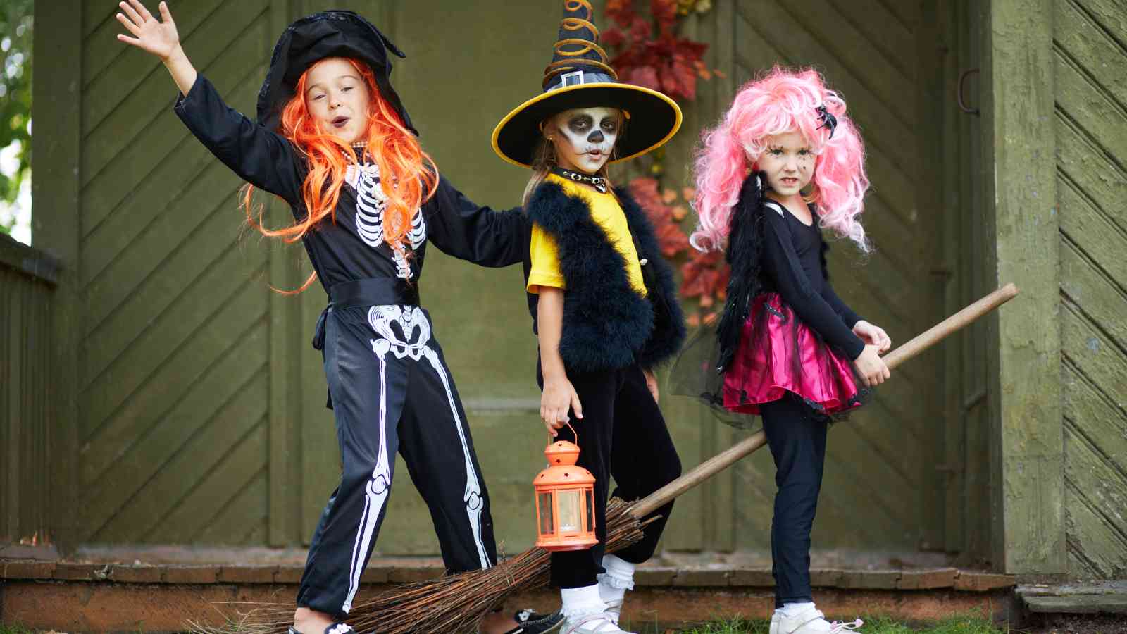 Three children in Halloween costumes pose outdoors; one dressed as a skeleton witch, one as a skeleton in a hat with a lantern, and one in a pink wig and dress holding a broom.