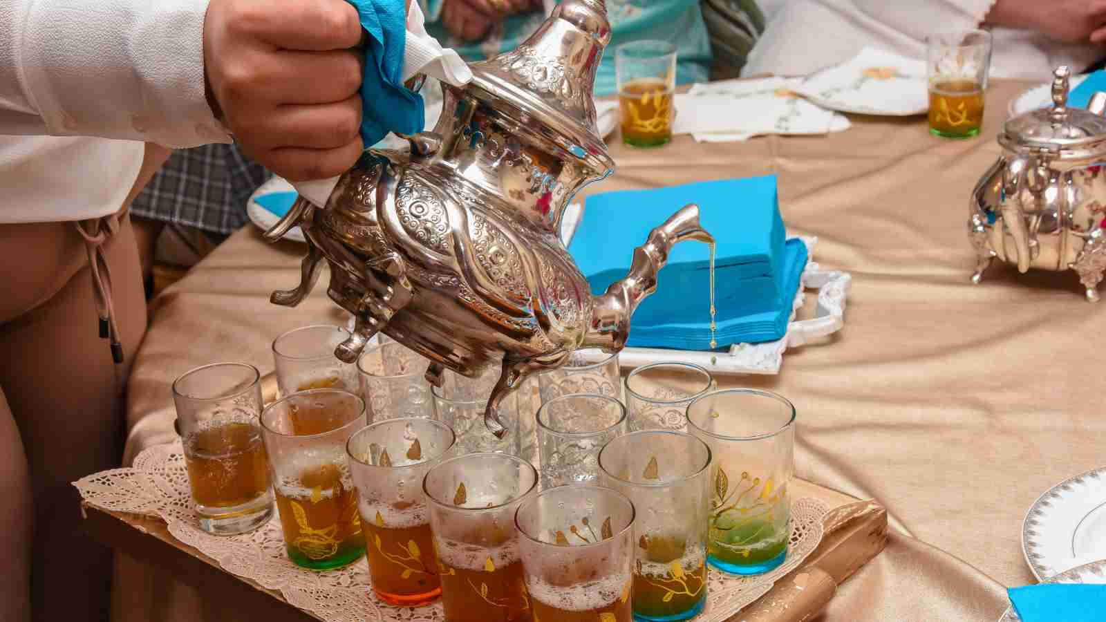A person pours tea from an ornate silver teapot into a row of decorated glasses on a table set with napkins and plates.