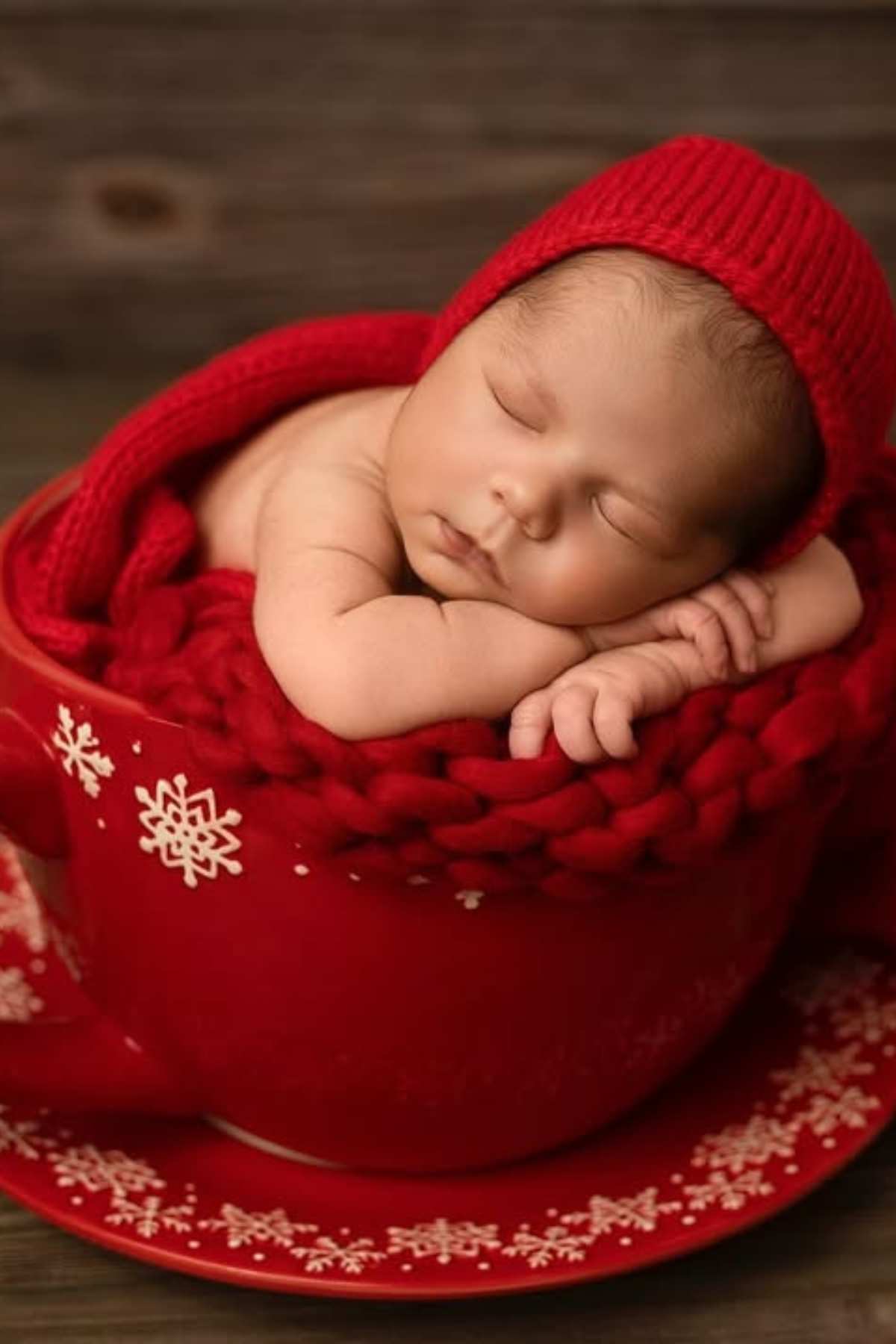 A newborn baby wearing a red knit hat sleeps in a large red teacup with snowflake designs, resting on a matching saucer.