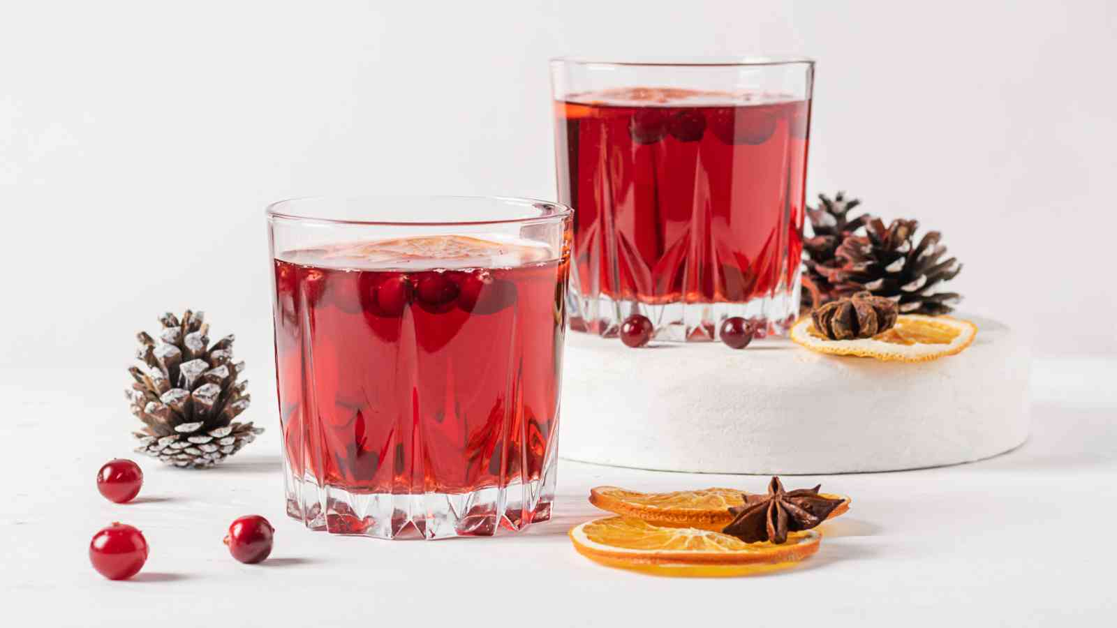 Two glasses of red beverage with cranberries, surrounded by dried orange slices, star anise, cranberries, and pinecones on a white surface.