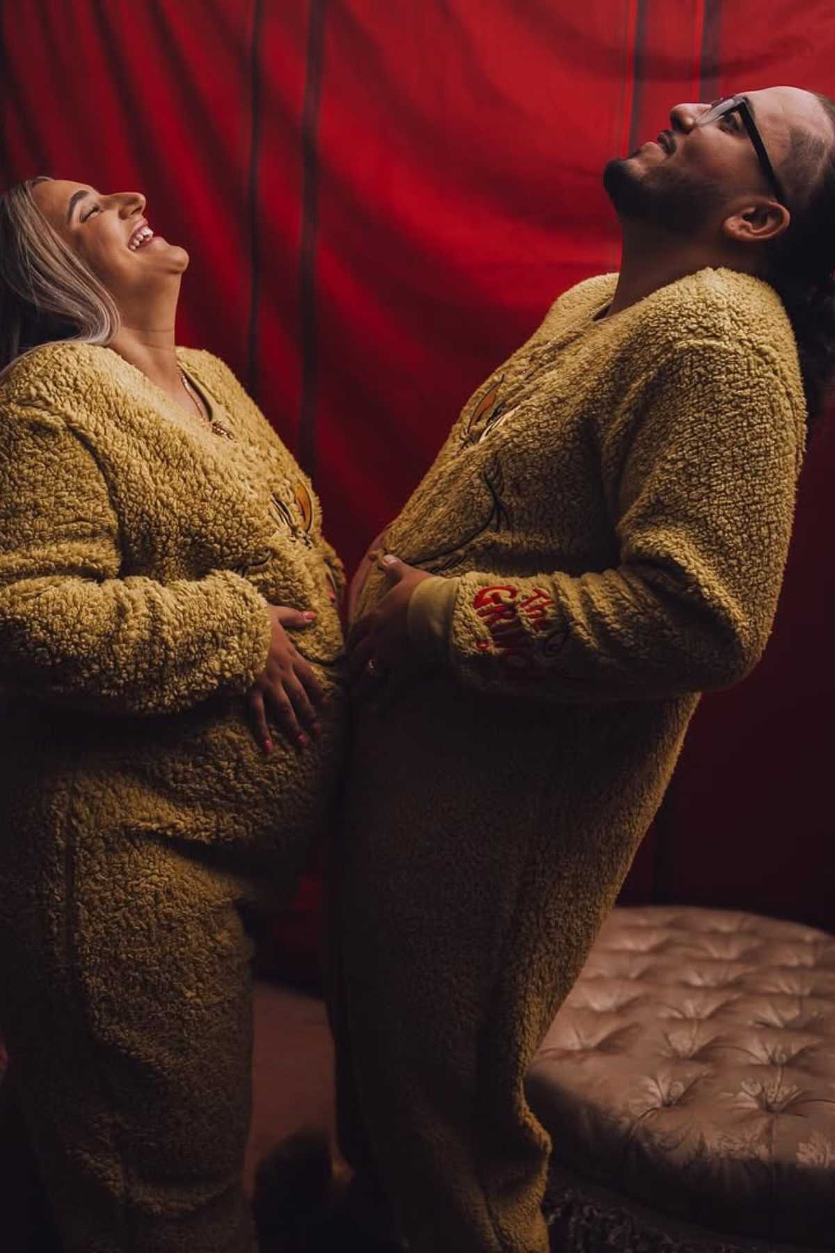 Two people in matching yellow fuzzy onesies stand side by side, both holding their stomachs and laughing, in front of a red backdrop.