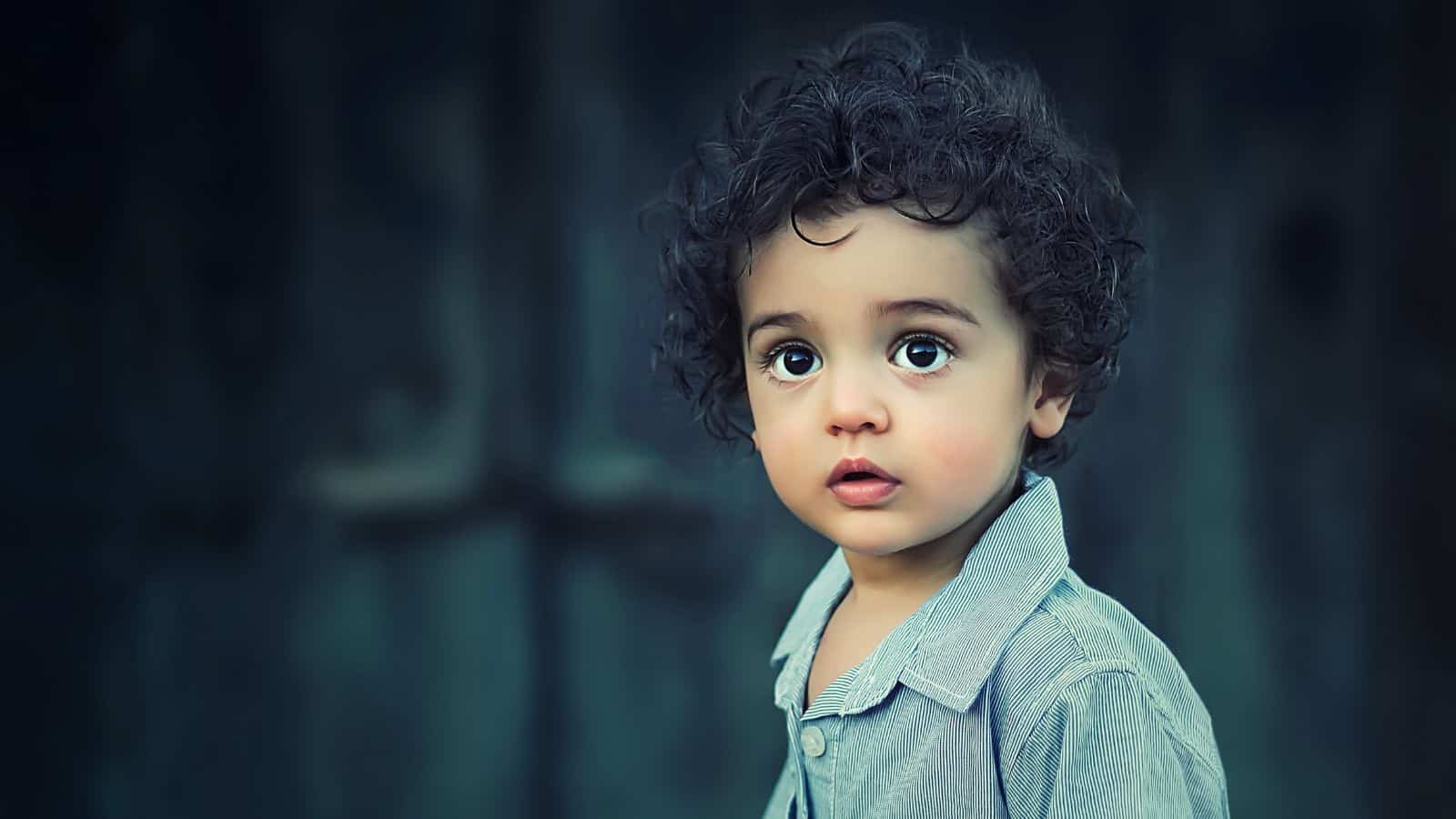 A young child with curly hair and large eyes looks slightly to the side, wearing a light gray shirt with a dark, blurred background.