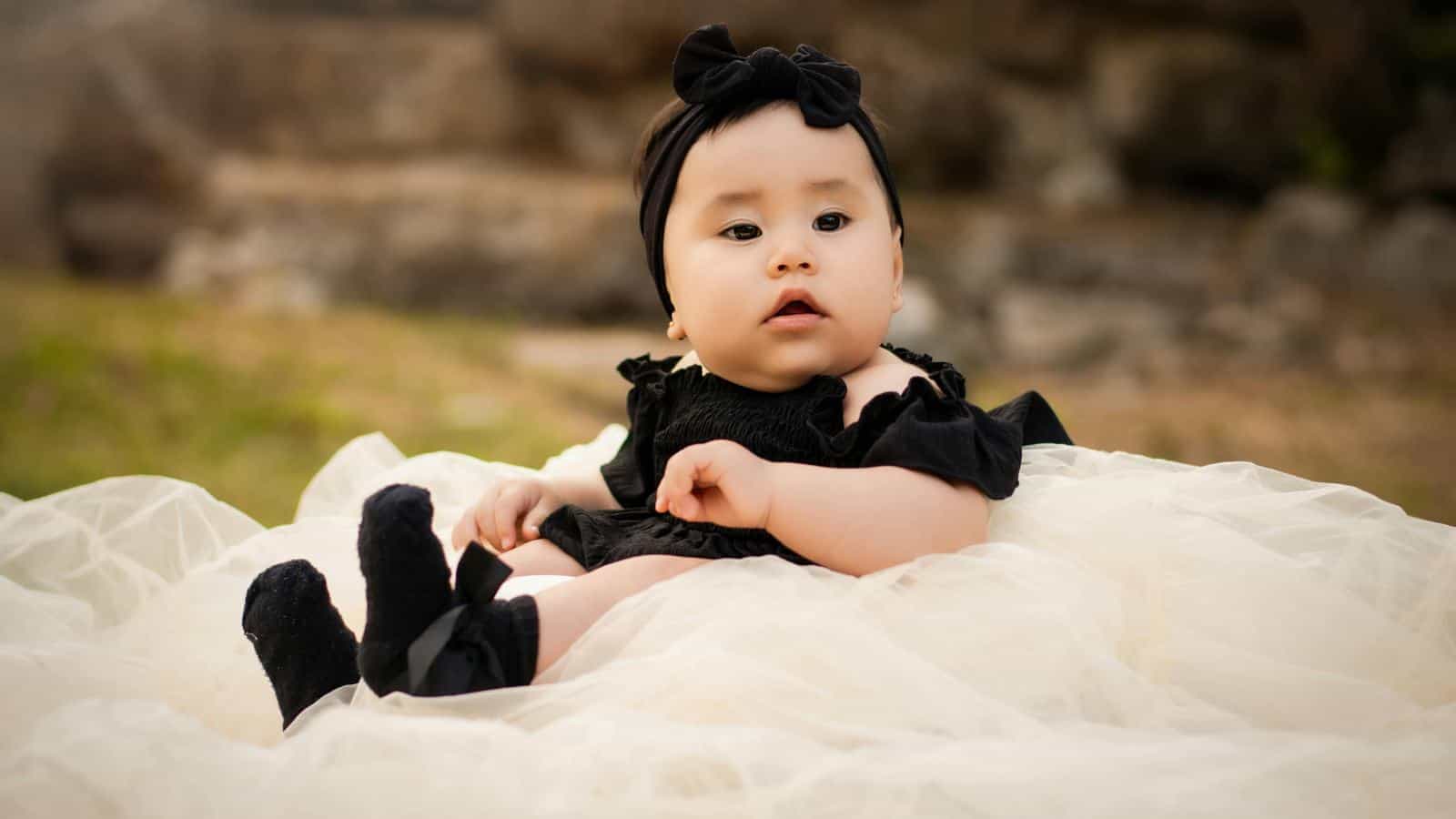 A baby wearing a black headband, black dress, and black socks sits on a cream-colored tulle fabric outdoors.