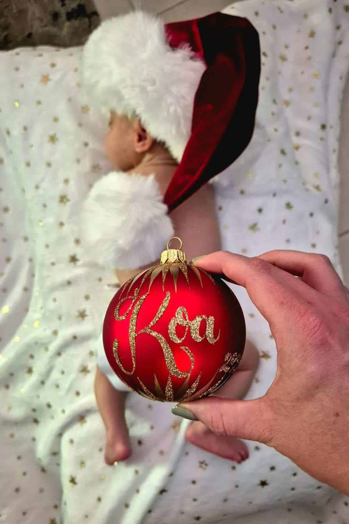 A baby in a Santa hat lies on a star-patterned blanket; a hand holds a red ornament with "Koa" written on it in the foreground.