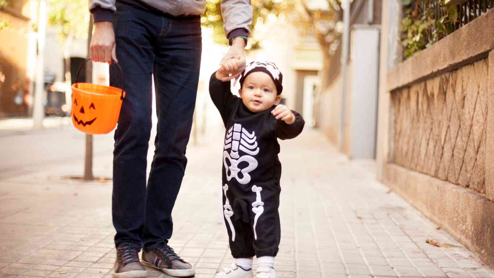 An adult holds a toddler's hand while walking outdoors; the toddler wears a skeleton costume and the adult carries an orange pumpkin-shaped bucket.