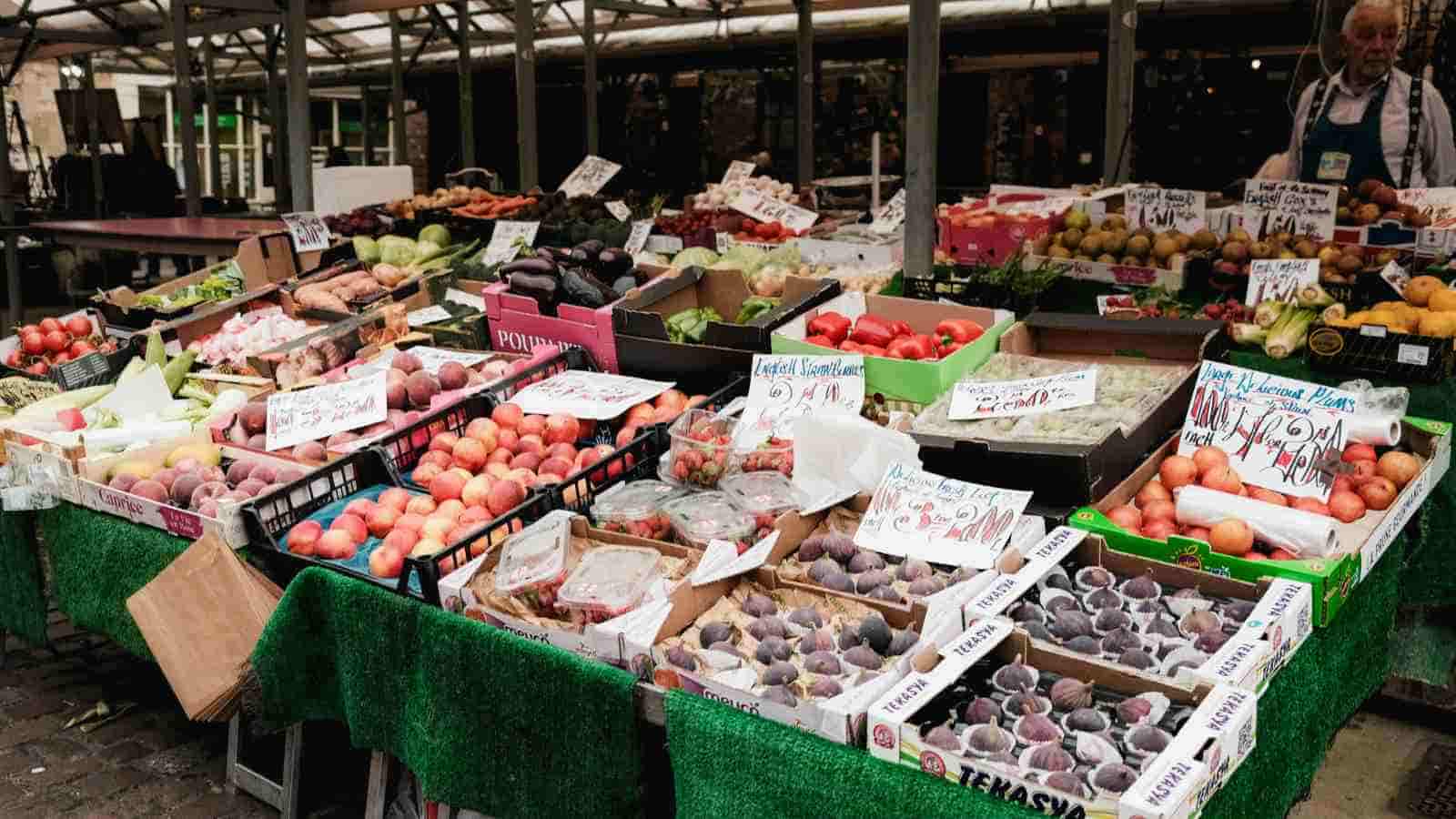 A market stall displays various fresh fruits and vegetables, including peaches, figs, and apples, with handwritten price signs on each box.