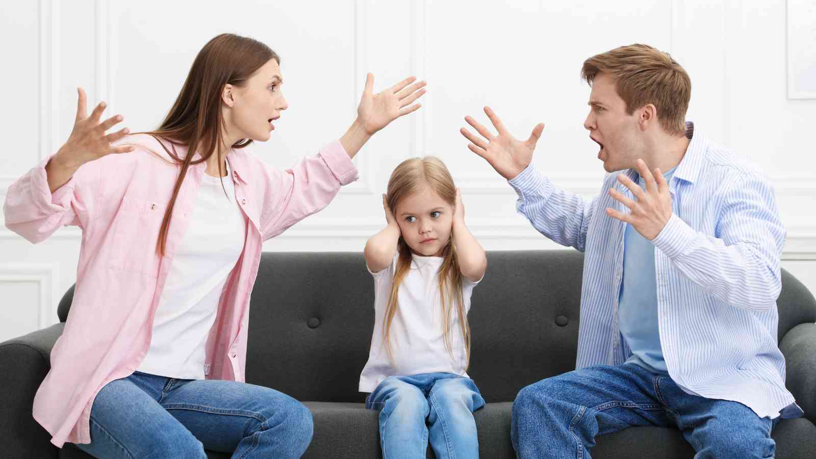 A woman and man are arguing on a couch while a young girl sits between them, covering her ears.