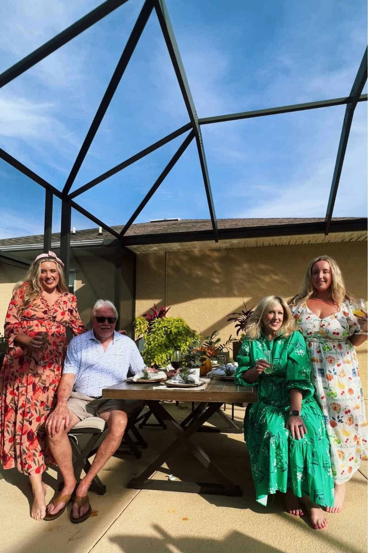 Four adults pose by an outdoor table set for a meal under a screened patio. Three women wear floral dresses; one man is seated wearing sunglasses and a short-sleeved shirt.