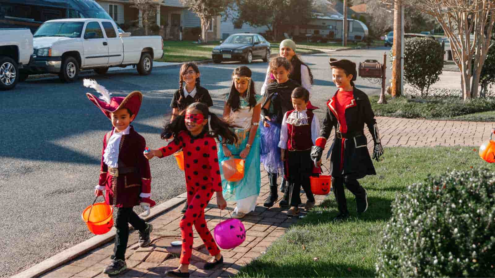 A group of children in various costumes walk on a sidewalk with orange buckets, likely trick-or-treating in a suburban neighborhood.