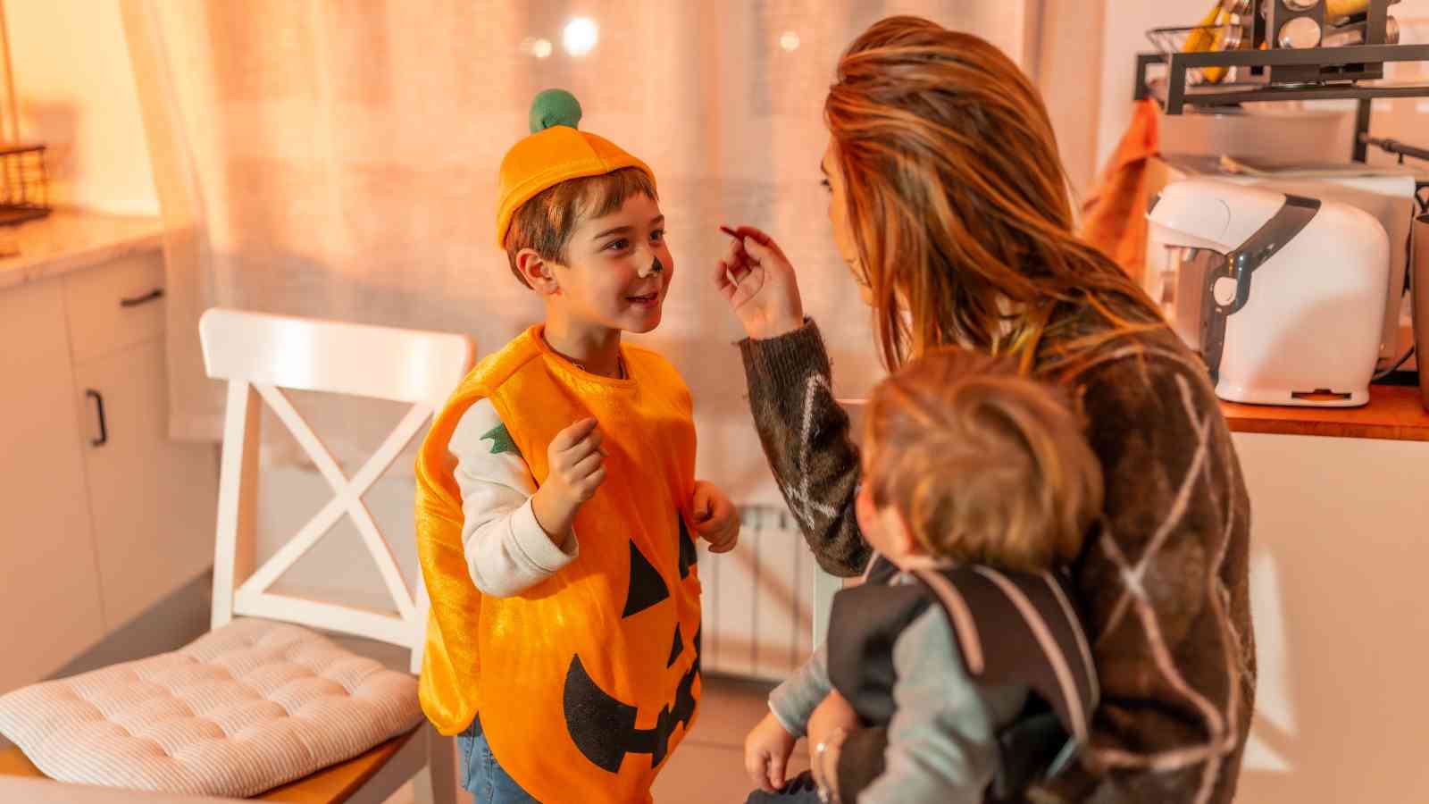 A woman holds a toddler while talking to a boy dressed in a pumpkin costume in a kitchen.