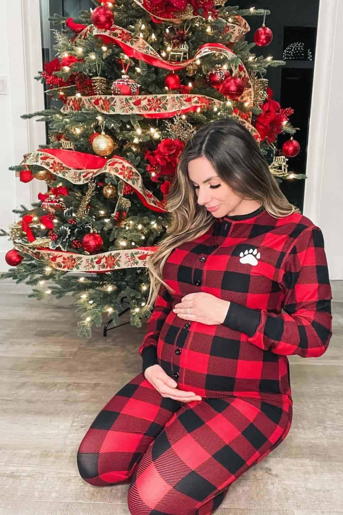 A woman in red and black plaid pajamas kneels in front of a decorated Christmas tree, cradling her pregnant belly and looking down.