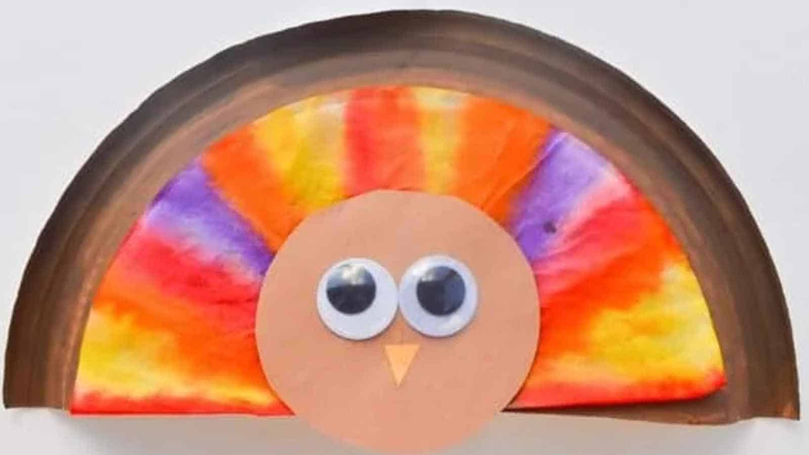 A paper plate turkey craft with a colorful, tie-dyed tail, brown border, and a round face with two large googly eyes and a small orange beak.