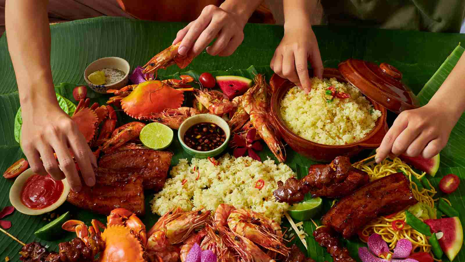 Three people share a meal of grilled seafood, rice, and vegetables, served on banana leaves with various sauces, as they eat with their hands.