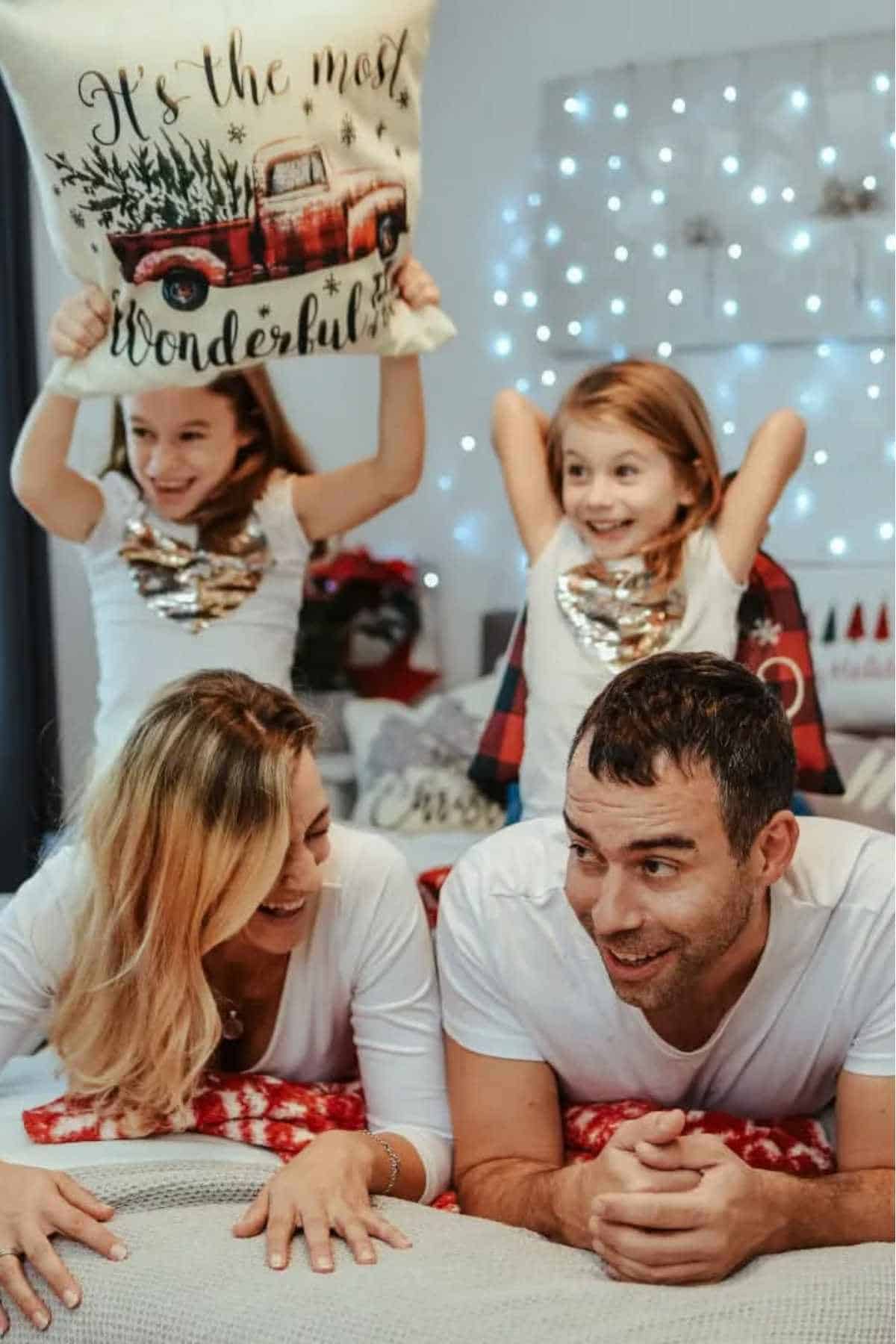 A family of four in white shirts smiling and relaxing indoors; one child holds a festive pillow, with string lights and holiday decorations in the background.