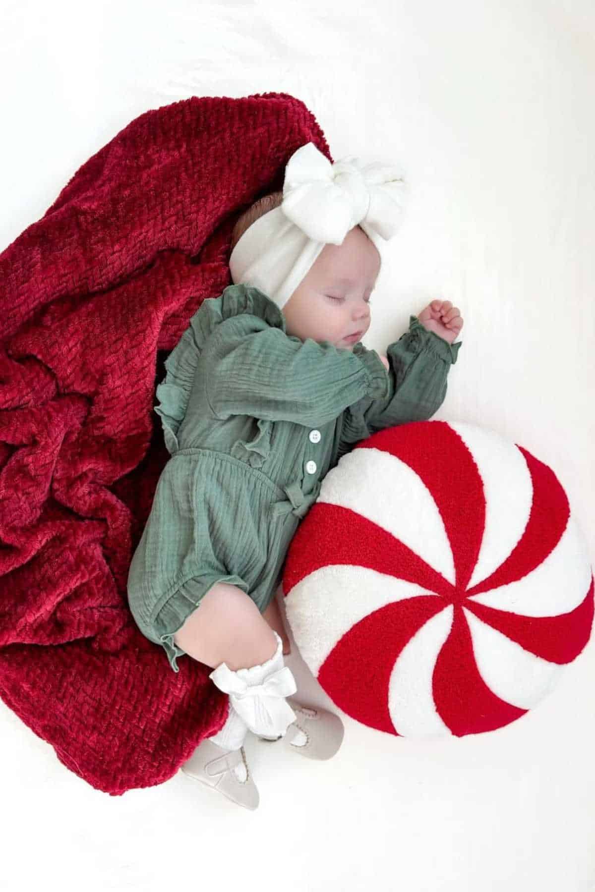 A baby in a green outfit and white headband sleeps on a red blanket, next to a round cushion with a red and white peppermint swirl pattern.