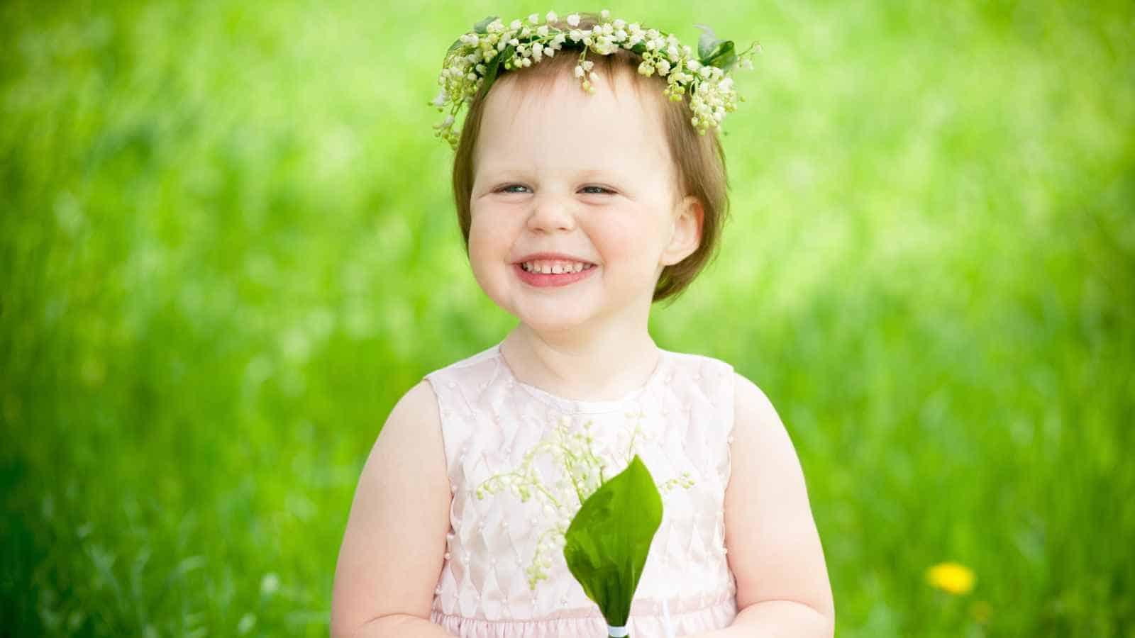A young child wearing a flower crown and light pink dress smiles while holding a green leaf, standing outdoors in a grassy area.