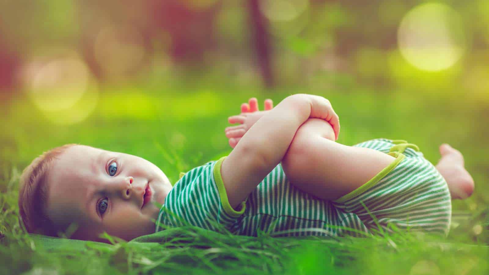 A baby in a green striped outfit lies on the grass outdoors, holding their feet with both hands and looking toward the camera.