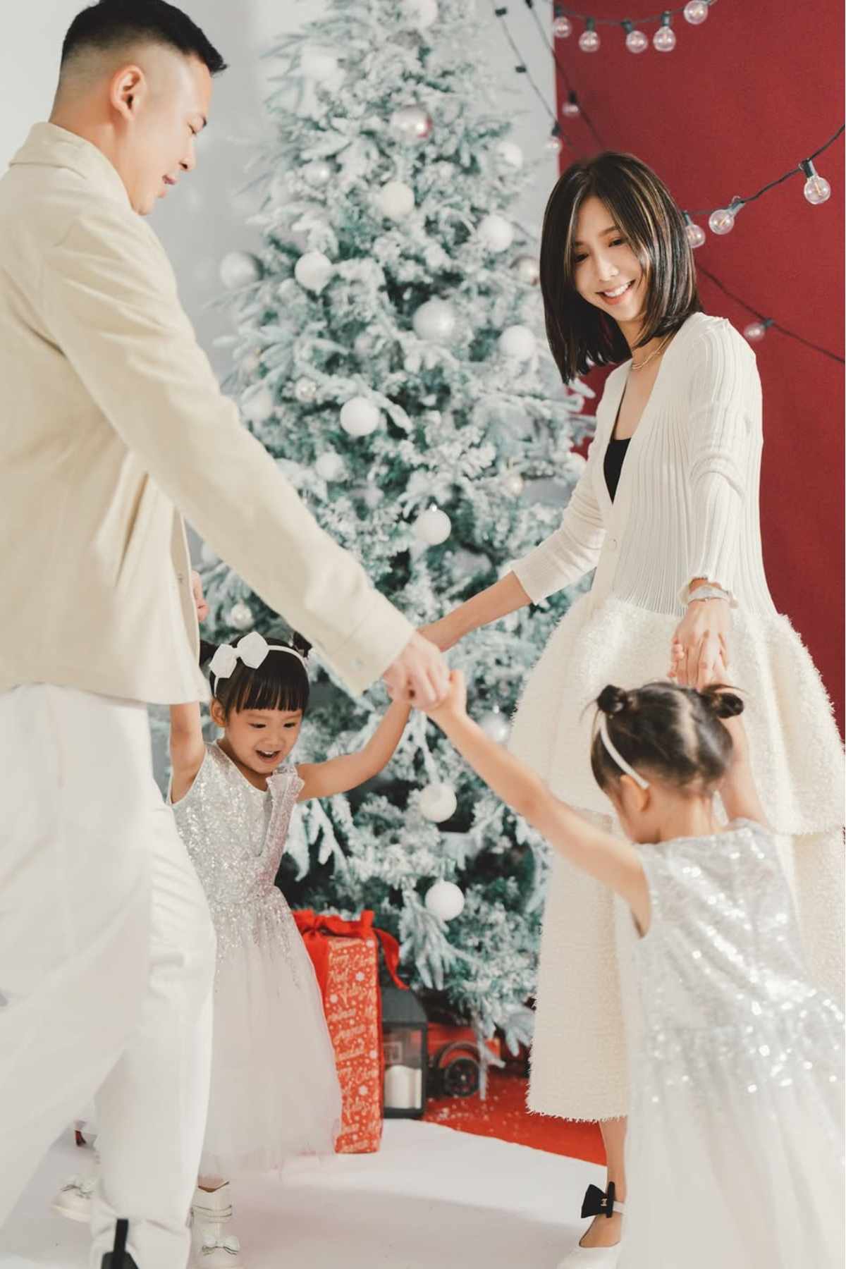 Two adults and two children hold hands in a circle and smile in front of a decorated Christmas tree with gifts and string lights.