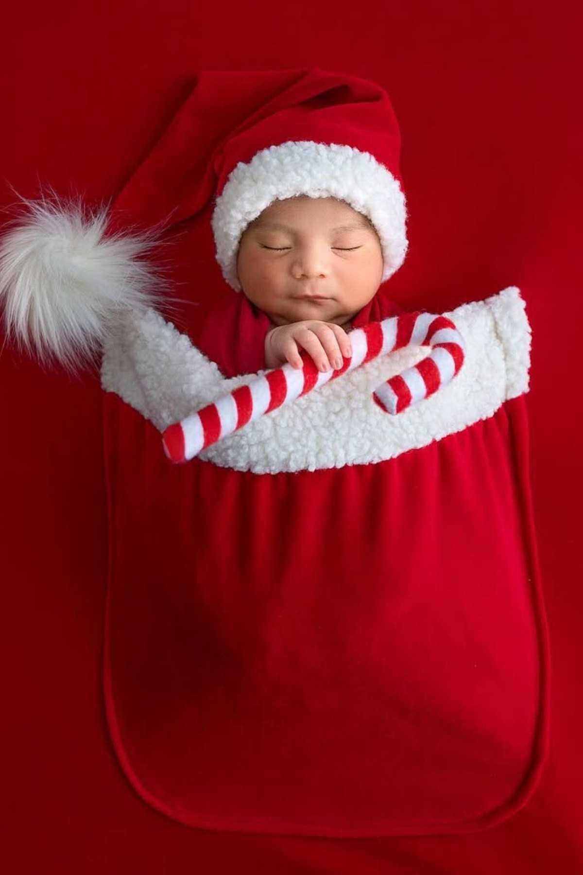 Newborn baby dressed in a Santa hat, lying in a red Christmas stocking with a white trim and holding a striped candy cane prop, on a red background.