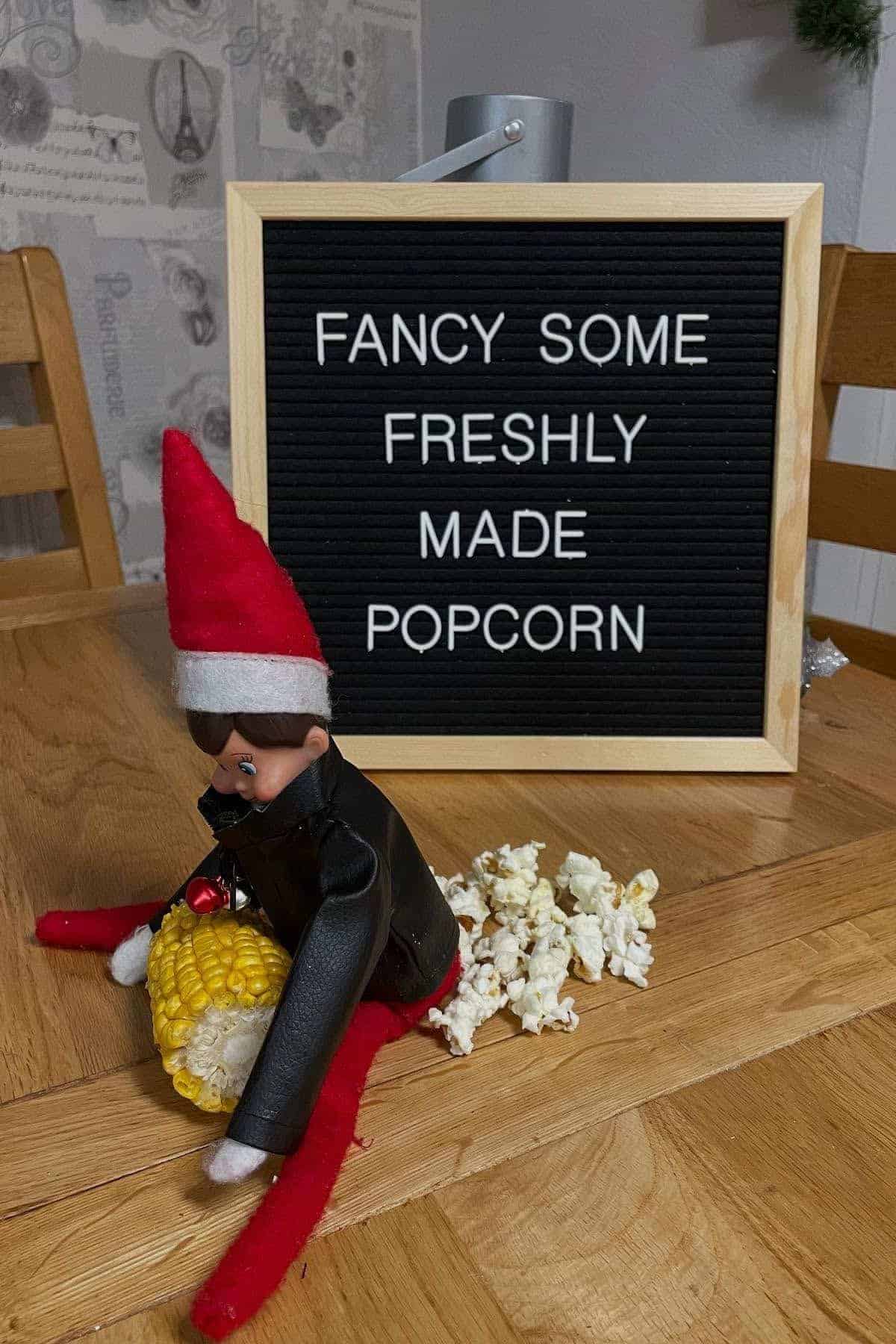 A toy elf sits on a table holding a corn cob with popped popcorn scattered behind it. A letter board in the background reads "Fancy some freshly made popcorn.
