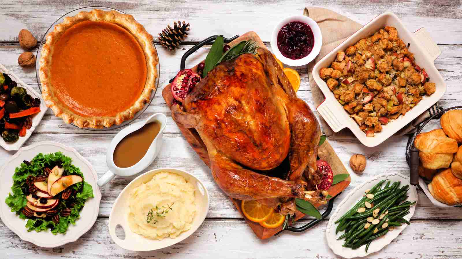 A roasted turkey surrounded by mashed potatoes, stuffing, green beans, salad, pumpkin pie, gravy, cranberry sauce, and bread rolls on a white wooden table.