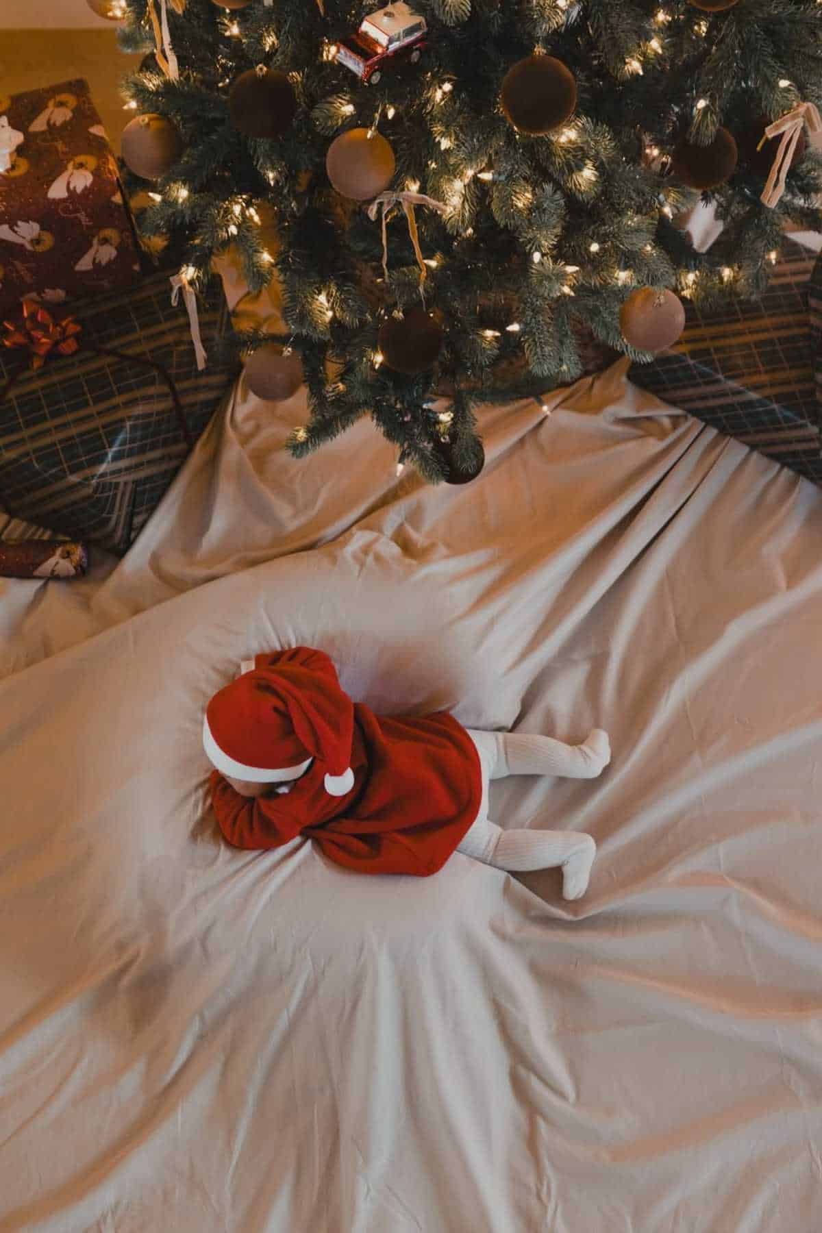 A baby dressed in a red Santa outfit lies on a white blanket under a decorated Christmas tree with wrapped gifts nearby.