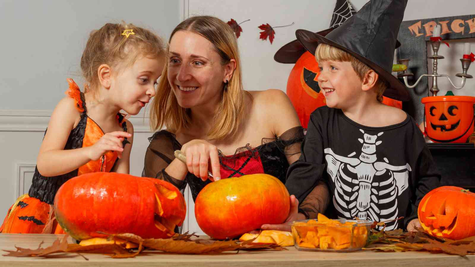 An adult and two children in Halloween costumes carve pumpkins together at a table decorated with autumn leaves and jack-o'-lanterns.