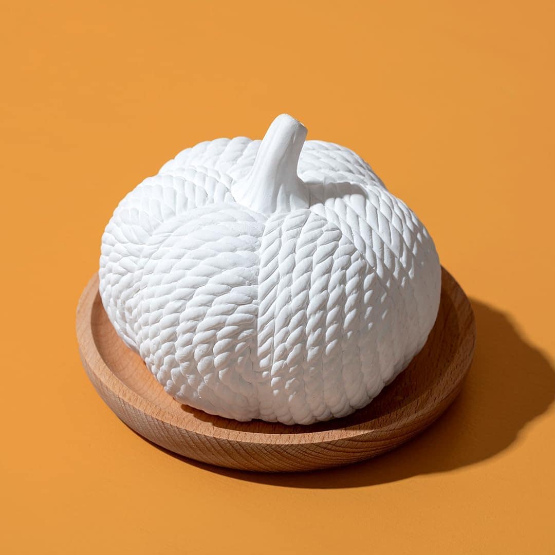 A white, textured ceramic pumpkin sits on a round wooden plate against an orange background.
