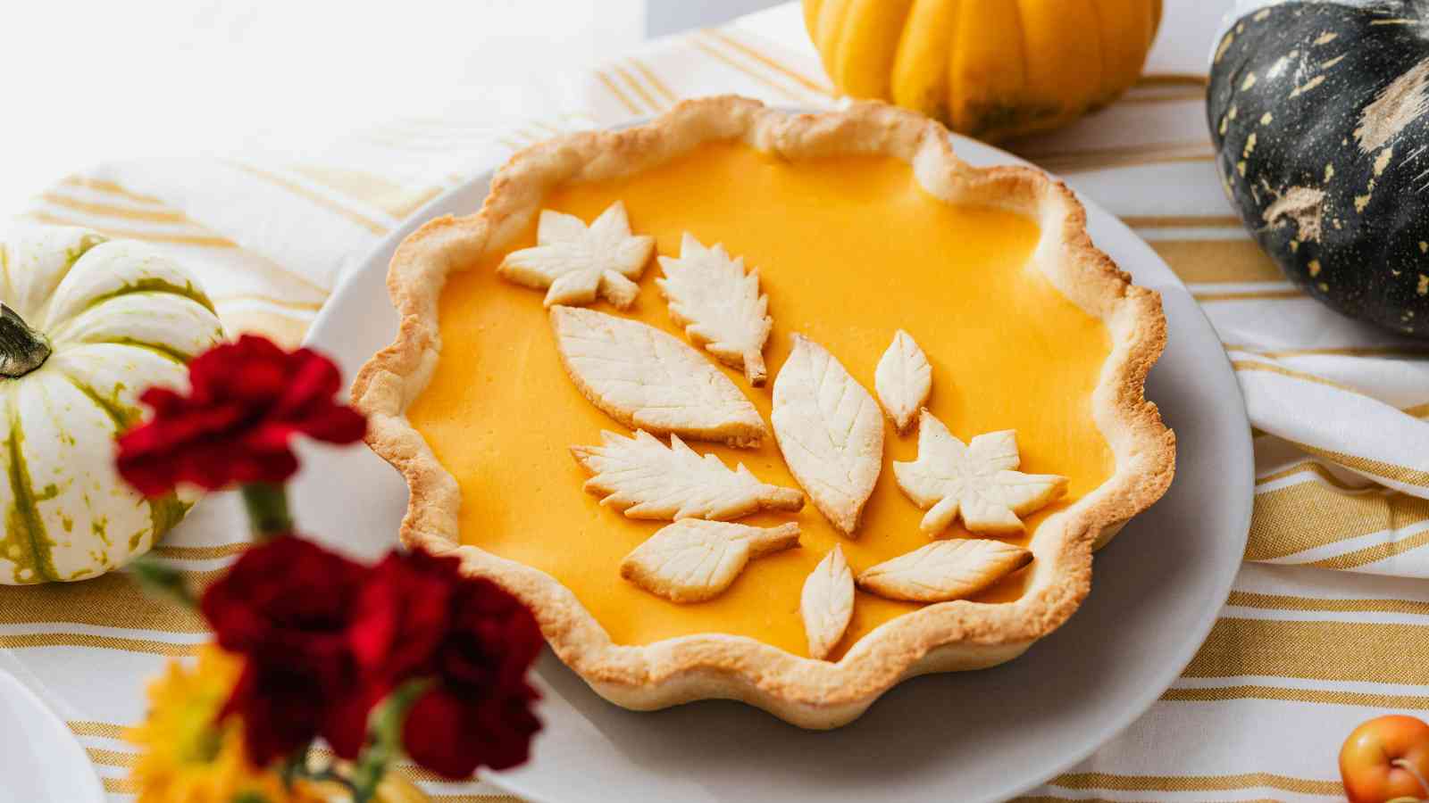 A pumpkin pie with decorative leaf-shaped crust pieces on top, placed on a white plate next to gourds and flowers.