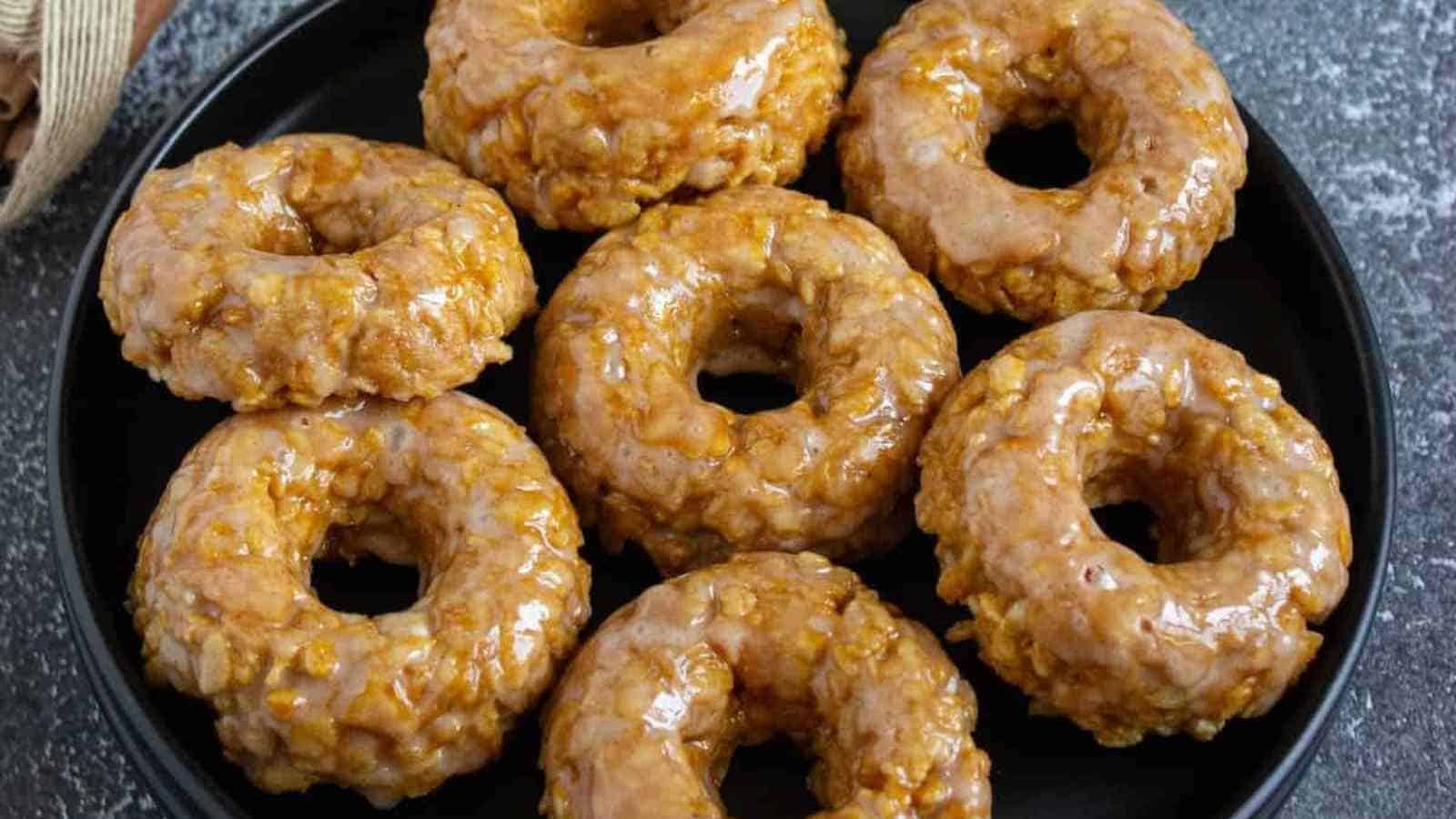 Eight glazed pumpkin Rice Krispie donuts are arranged on a black plate set on a dark textured surface.