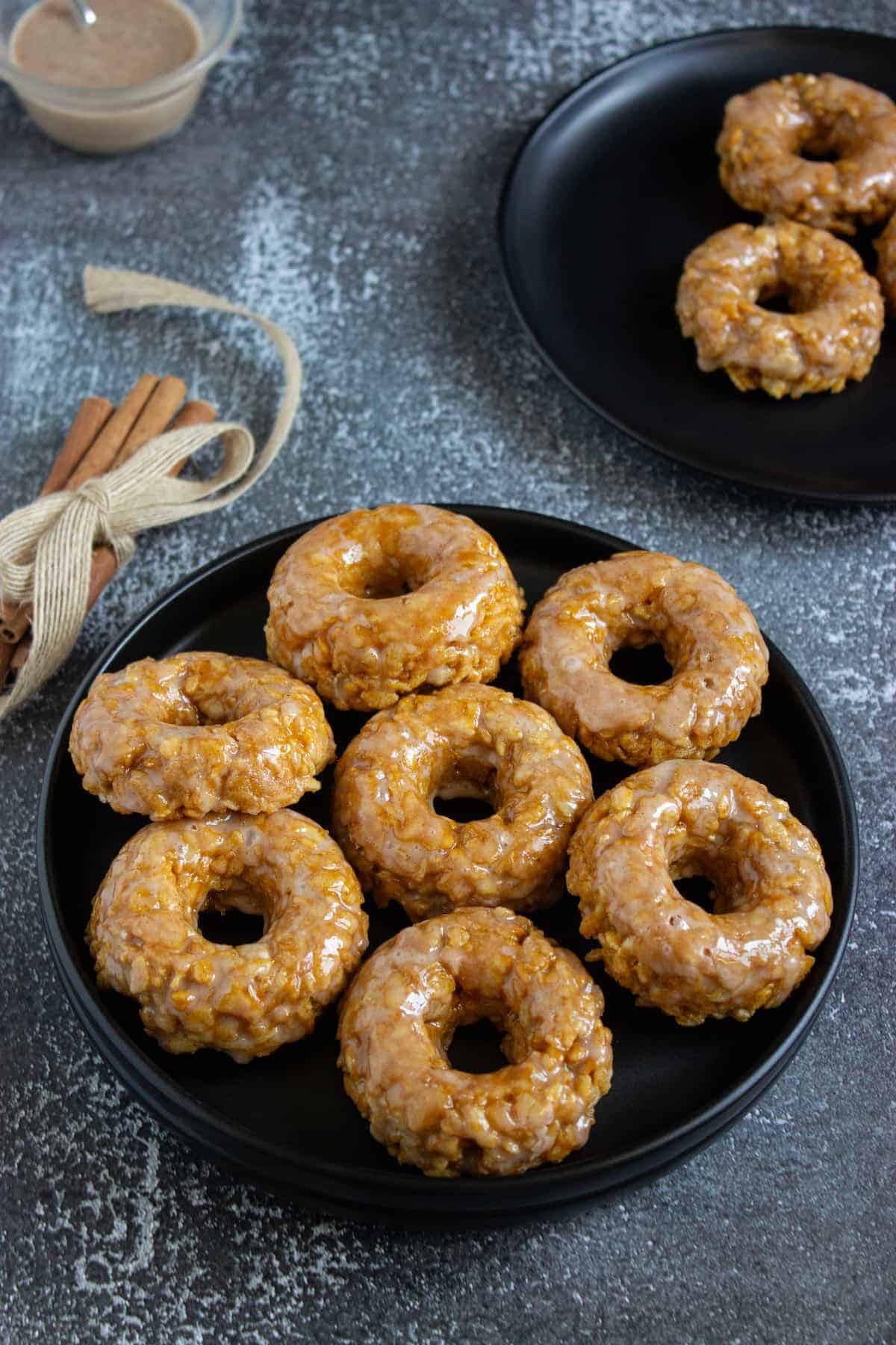 A black plate with eight glazed baked donuts sits on a textured surface; another plate with four donuts and a bundle of cinnamon sticks are nearby.