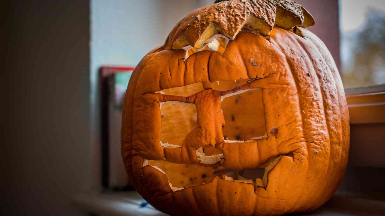 A carved pumpkin with a square-eyed face sits on a windowsill; its surface appears wrinkled and slightly decayed.