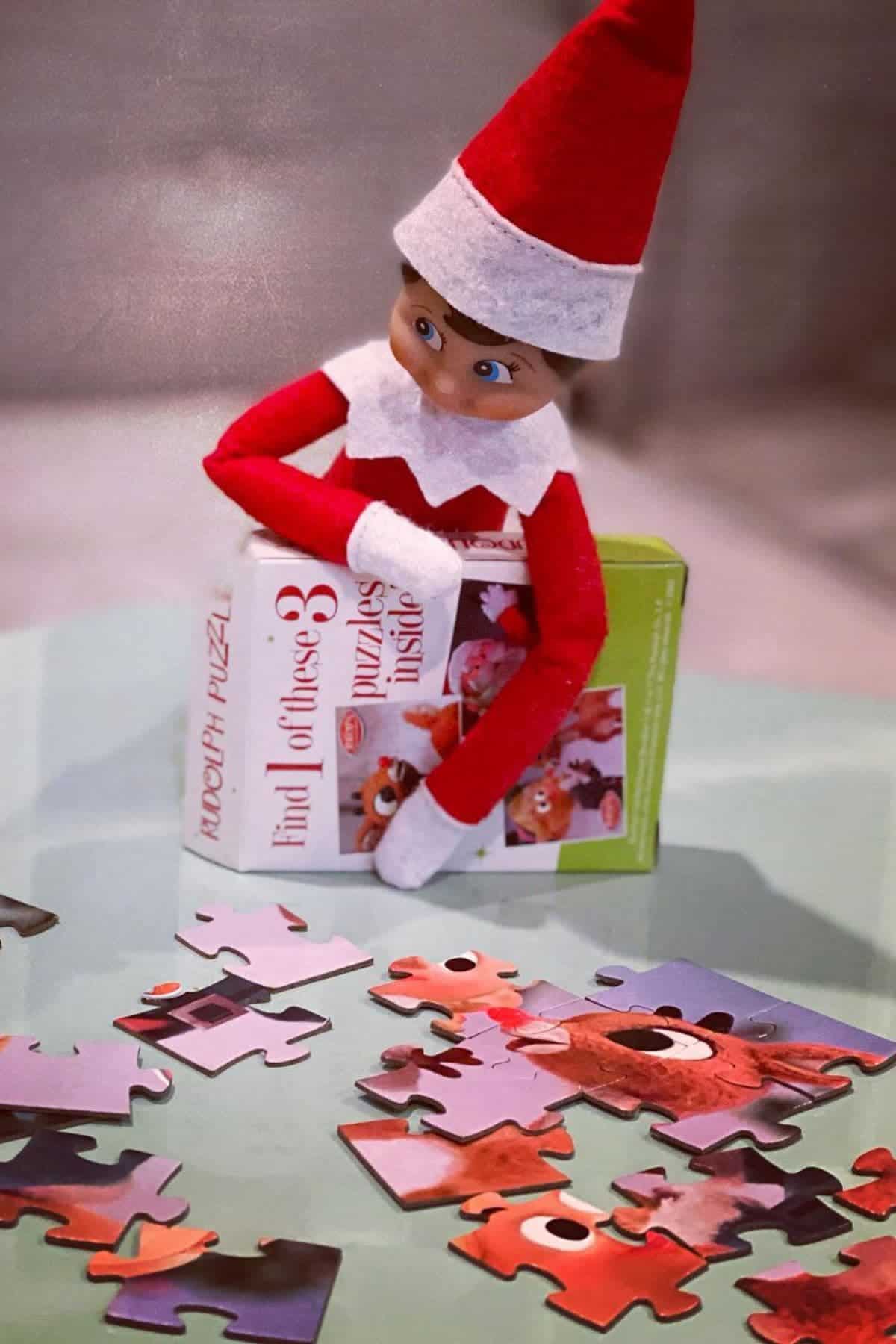 A toy elf in a red outfit sits behind a puzzle box, with several colorful puzzle pieces scattered on a glass table in front.