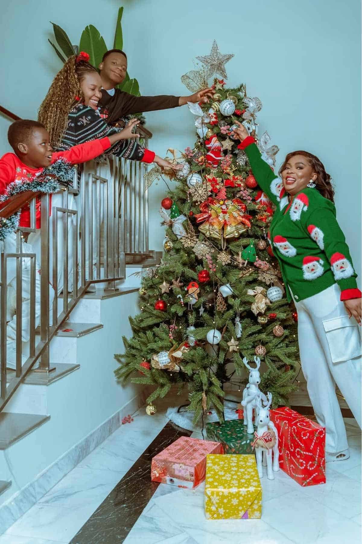 A family decorates a Christmas tree together; wrapped gifts sit underneath, and a woman places a star on top while others add ornaments.