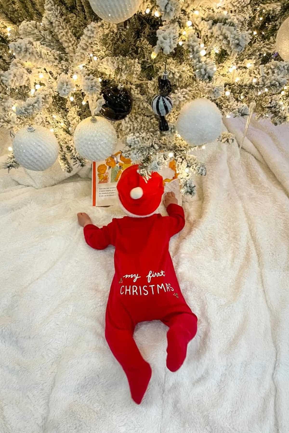 A baby in a red "My First Christmas" outfit lies on a white blanket under a decorated Christmas tree, looking at a book.