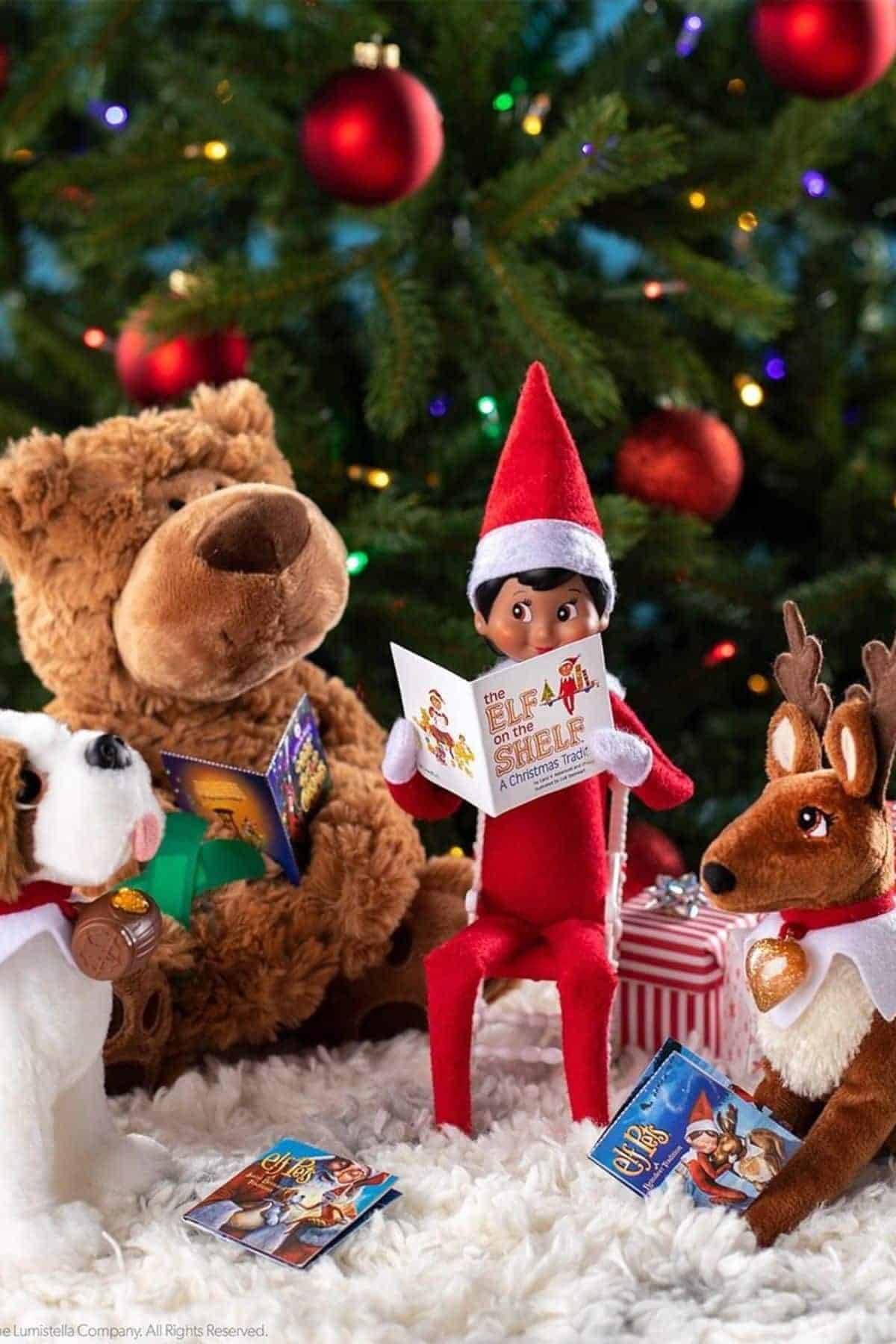 A plush elf doll sits reading a booklet, surrounded by a teddy bear, dog, and reindeer plush toys in front of a decorated Christmas tree with lights and gifts.