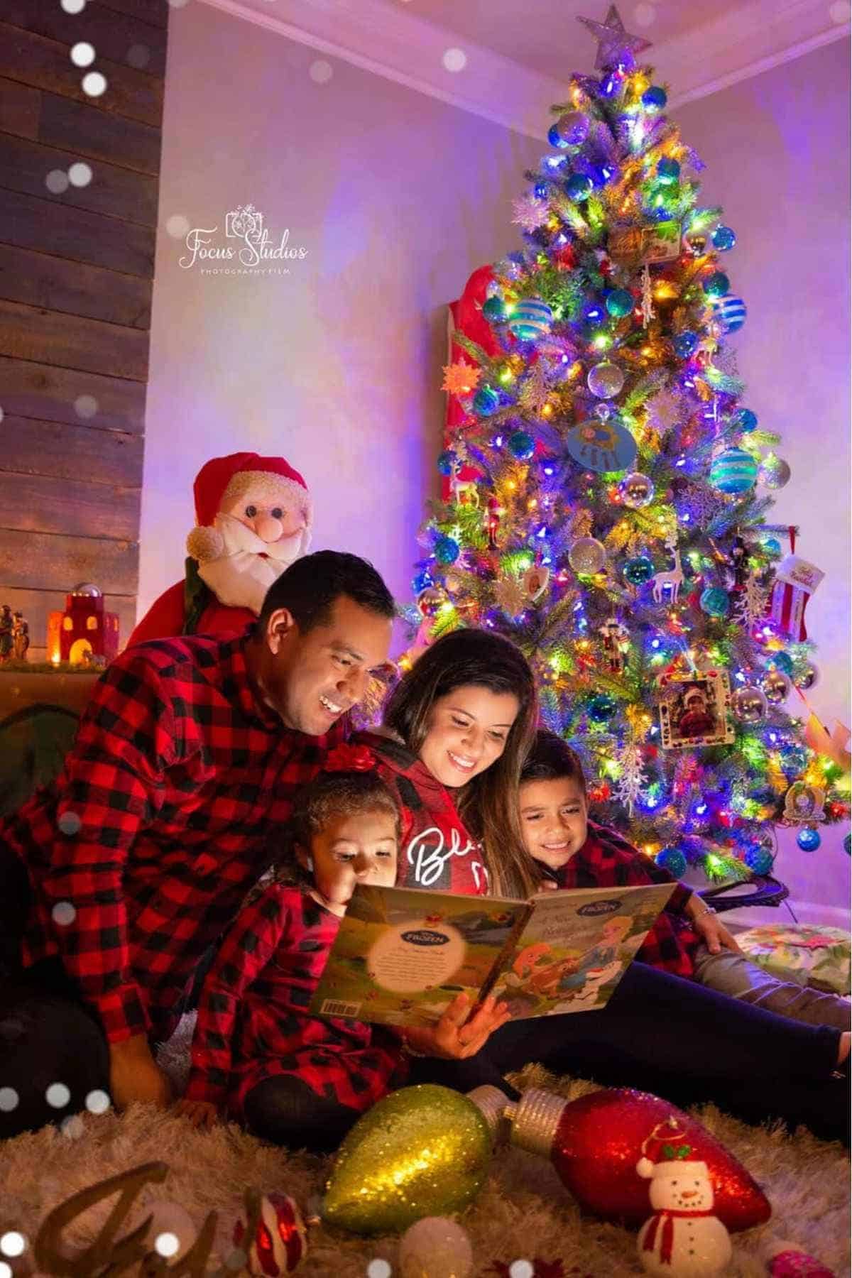 A family of four in matching red plaid shirts sits together on the floor by a decorated Christmas tree, reading a book in a cozy, festive room.