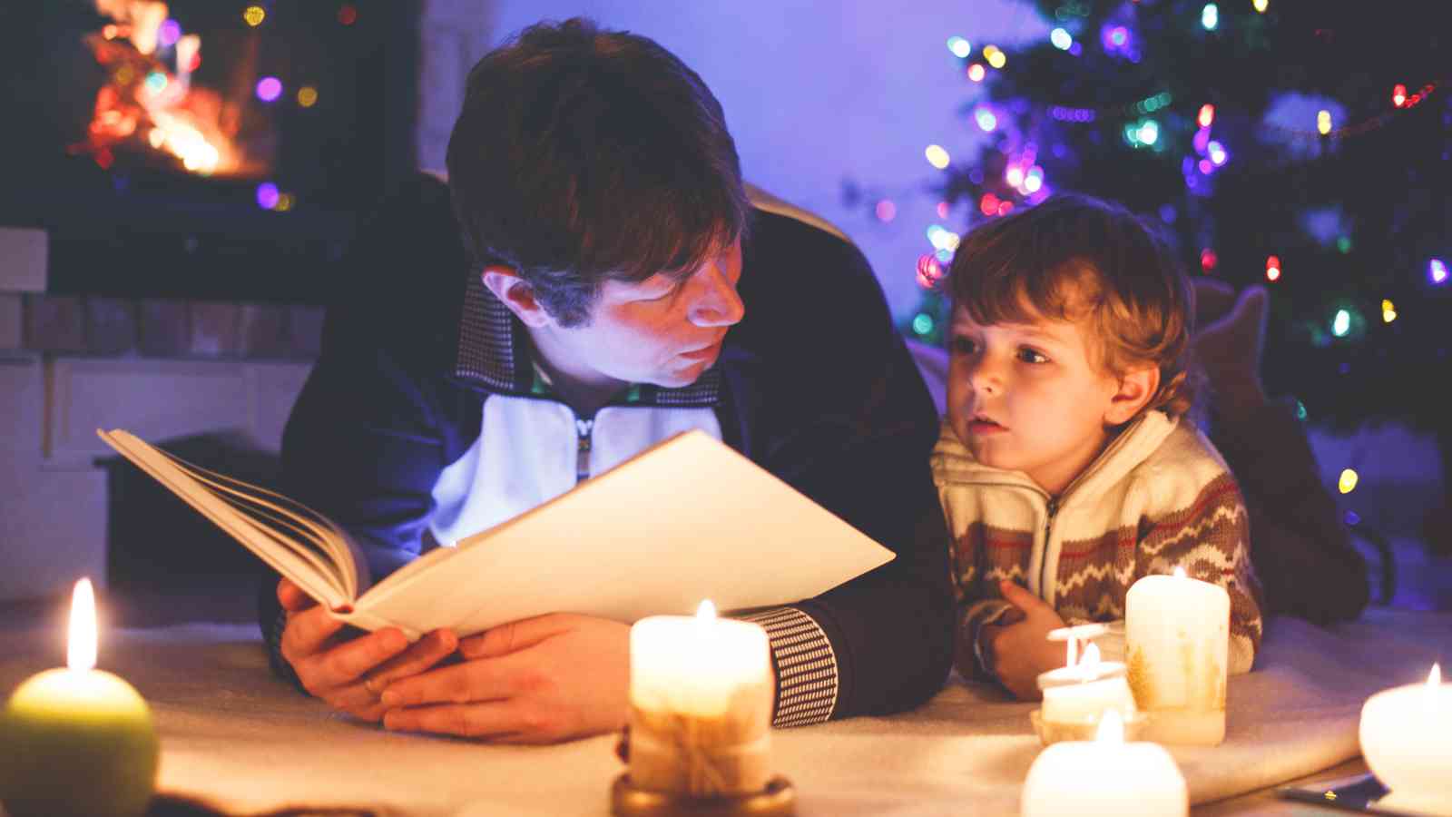 An adult and child lie on the floor surrounded by lit candles, reading a book together near a Christmas tree decorated with lights.